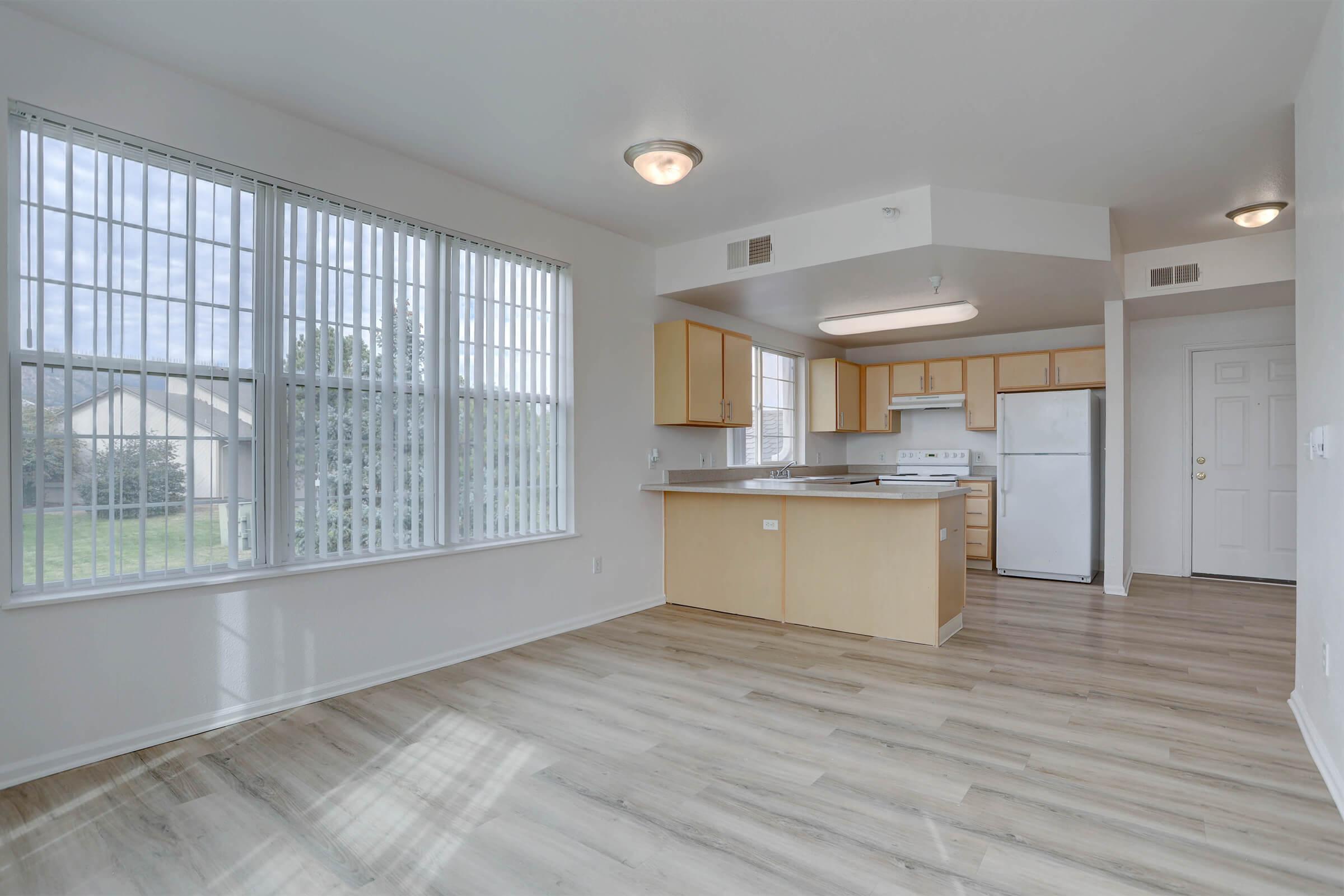 Interior view of a modern kitchen and living area featuring large windows with vertical blinds, light wood flooring, wooden cabinetry, a white refrigerator, and an oven. The layout is open and bright, with natural light illuminating the space.