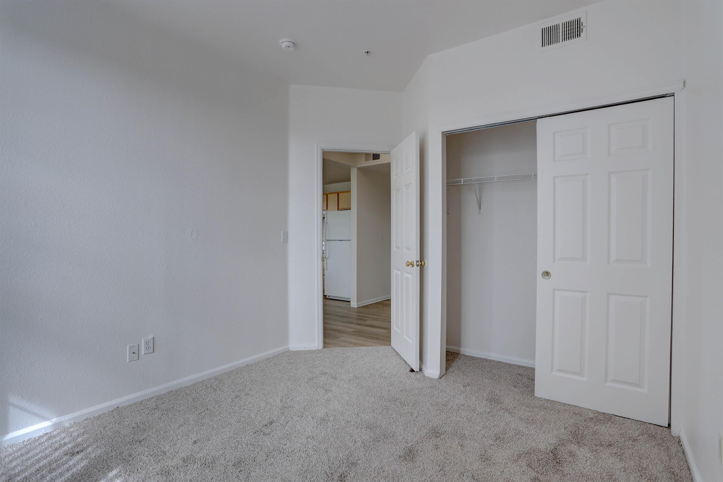 A small, empty room with light-colored walls and carpet. One side features a closed closet with double white doors, while another door leads to a kitchen area. Natural light illuminates the space, highlighting the clean and simple design.