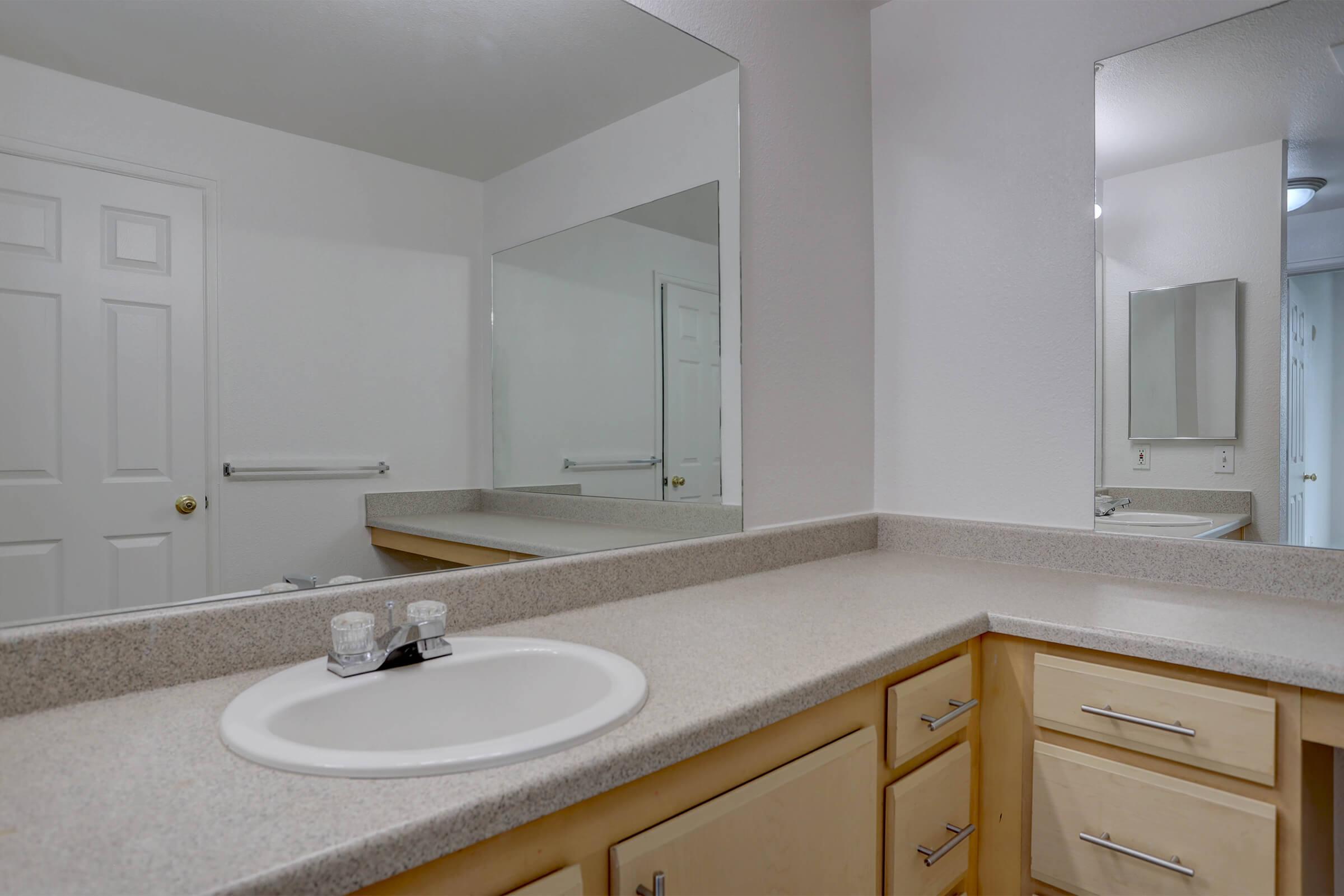 A clean and modern bathroom featuring a sink with a countertop, wooden cabinetry, and large mirrors. The walls are painted in a light color, and there is a towel bar visible. The overall design is sleek and minimalist, creating a bright and spacious feel.