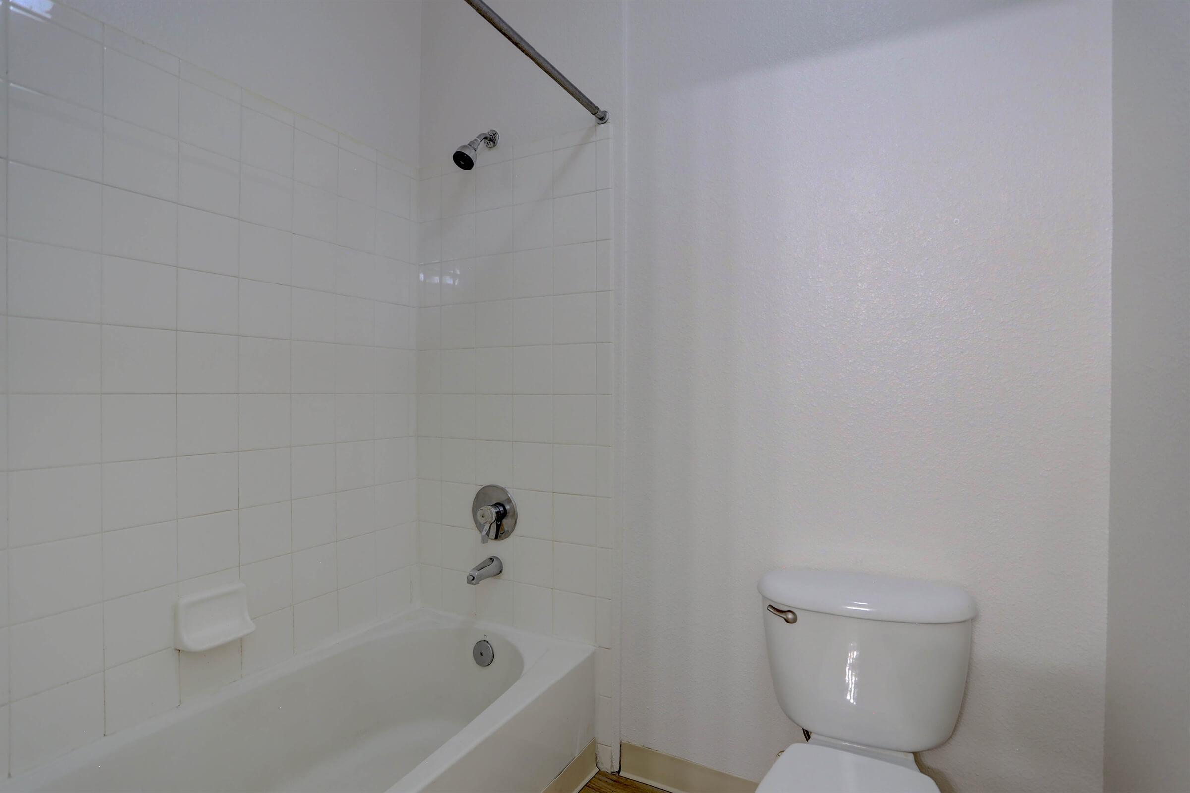 A simple bathroom featuring a white tiled shower area with a bathtub and an adjacent toilet. The walls and fixtures are clean and minimalistic, creating a bright and uncluttered space.