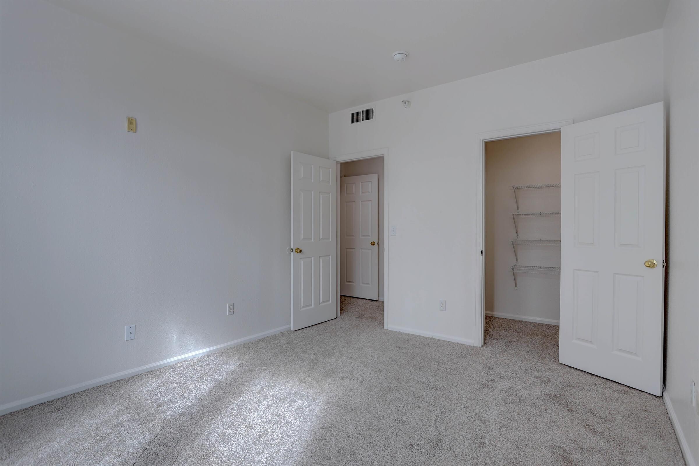 A clean, empty bedroom with light beige carpet and light beige walls. Two doors on the right lead to a closet and another room. Natural light enters from a window, creating a bright, open feel. The space is uncluttered, ready for personalization or furnishing.