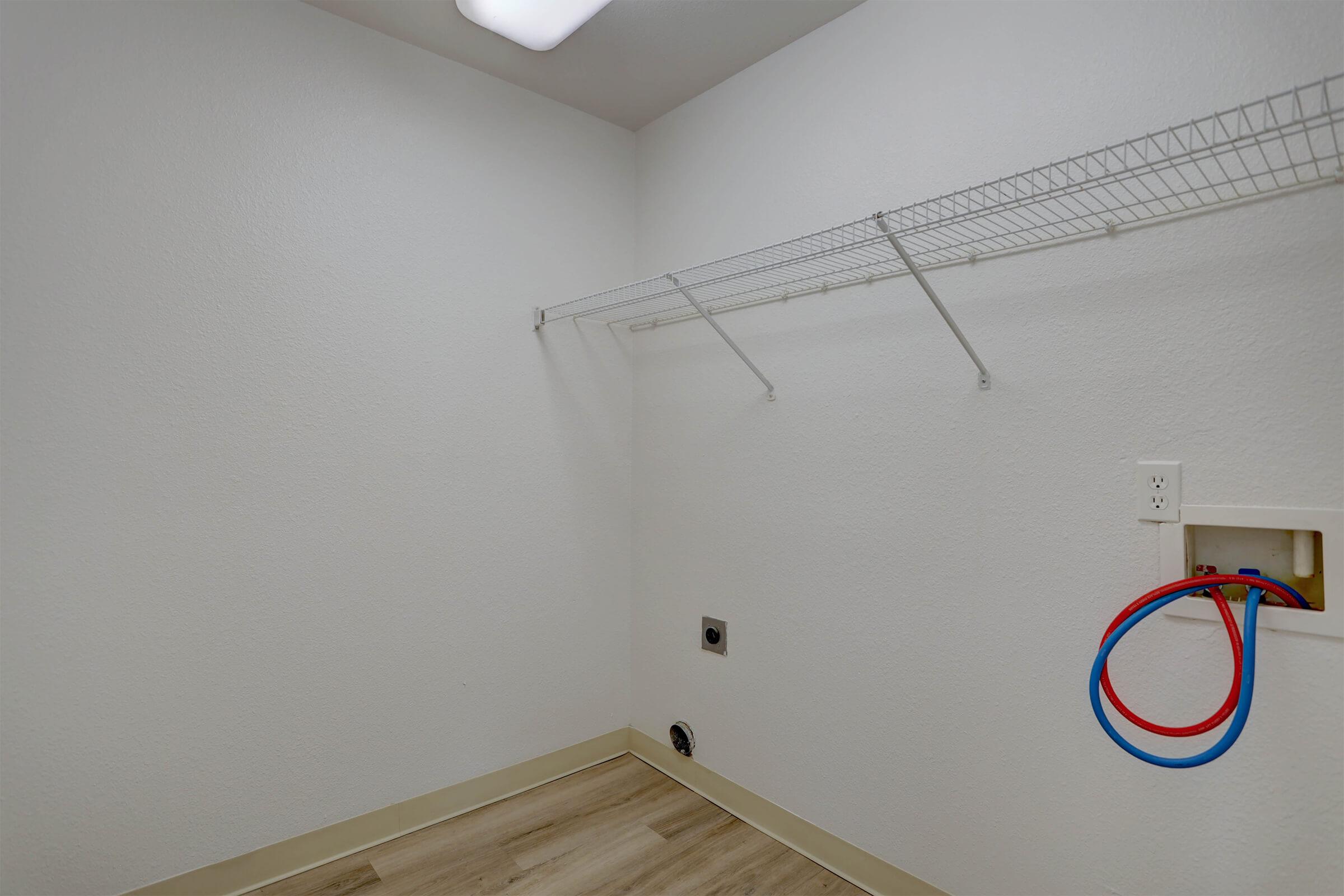 Empty laundry room with a white wall and a small section of shelving mounted on the wall. There is a power outlet and a blue and red hose coiled on the floor. The space features light-colored vinyl flooring and overhead lighting.