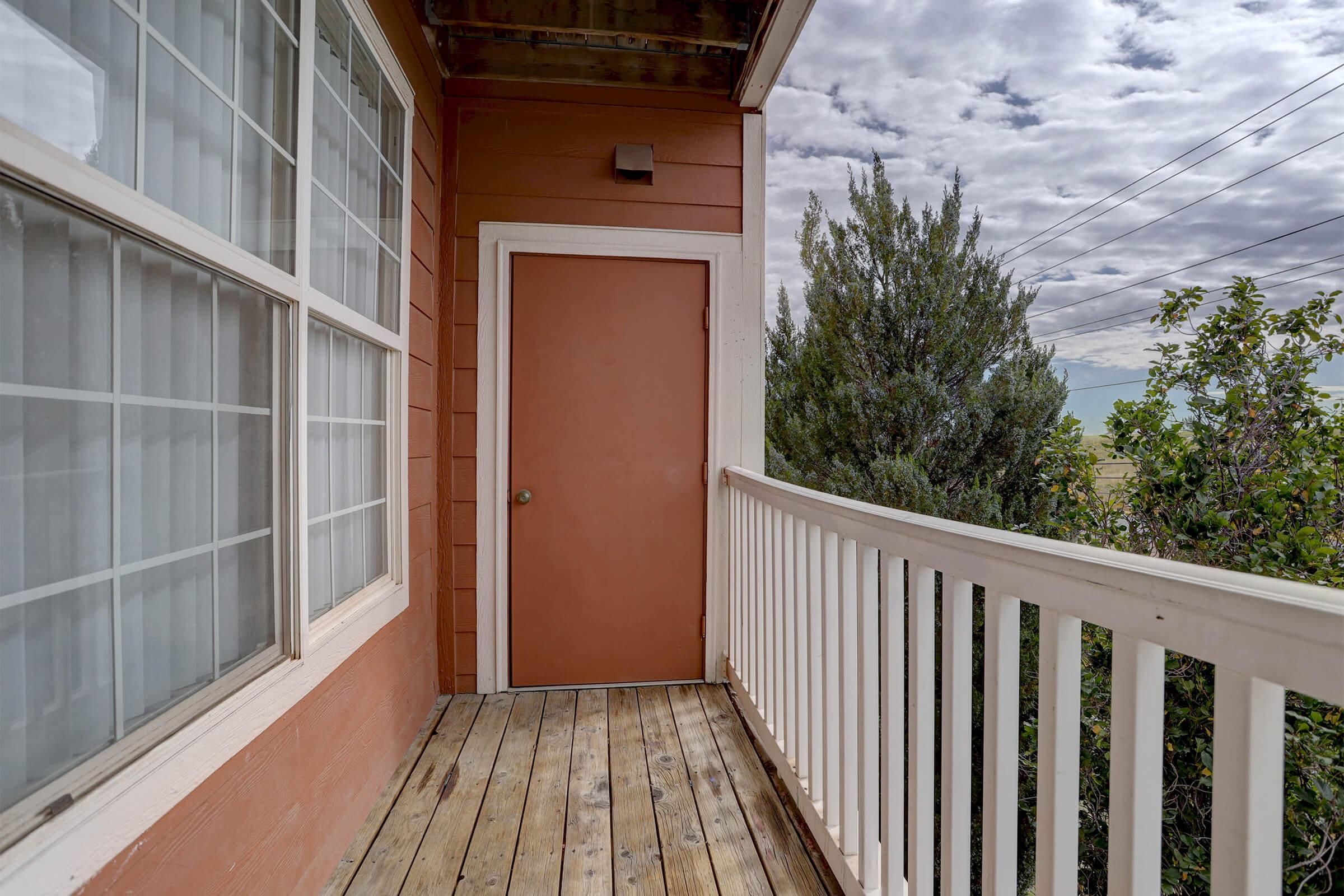 A small balcony with wooden flooring and a brown door. The wall is painted in orange, and there are large windows with white frames. In the background, green trees and a cloudy sky are visible, creating a serene outdoor atmosphere.