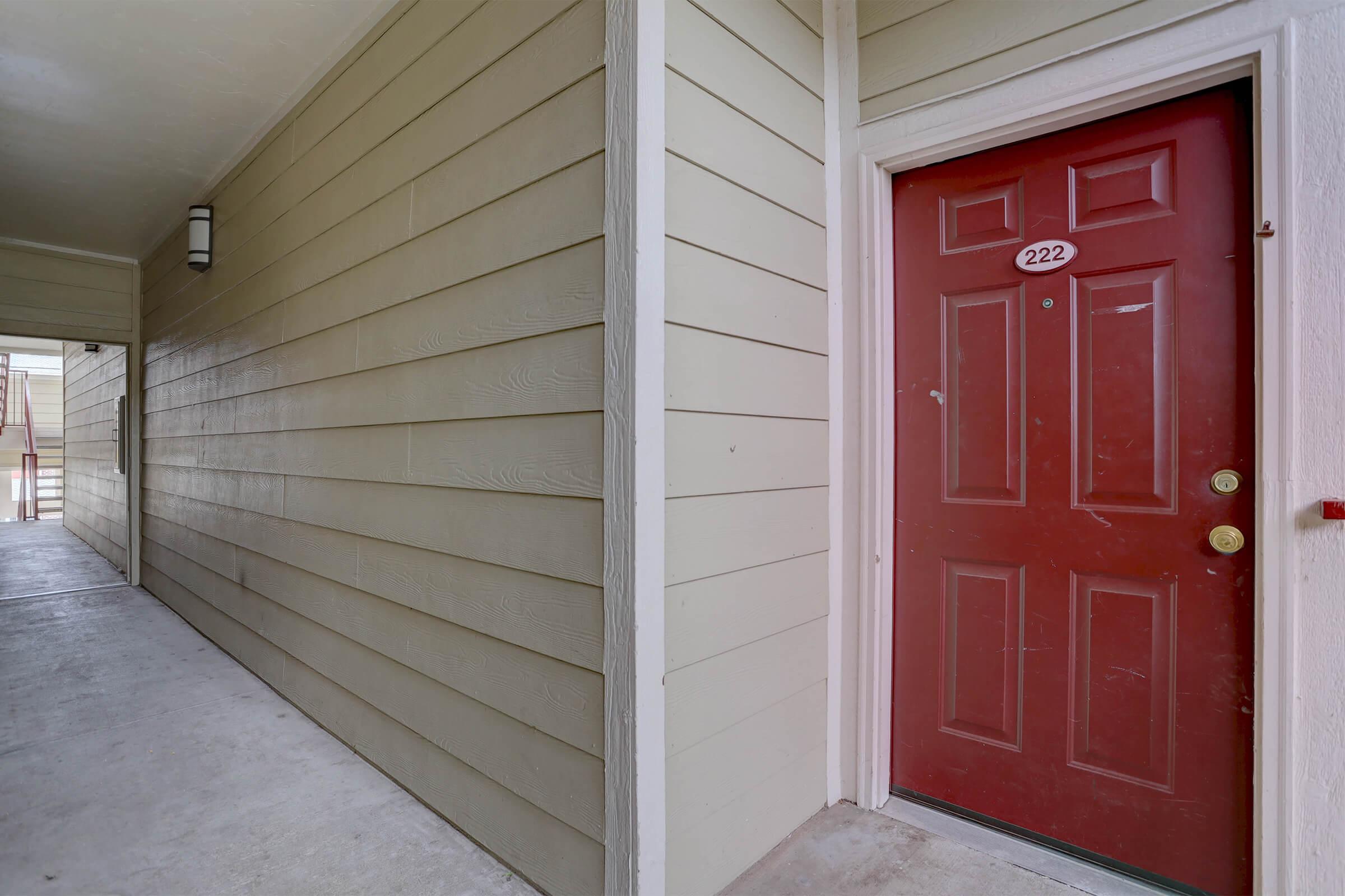 A close-up view of a red door numbered 222, set against a beige wall in a hallway. The hallway is well-lit and includes a lamp fixture on the wall. The floor is concrete, and there is a visible corridor leading to another entrance. The entrance is part of a multi-unit residential building.