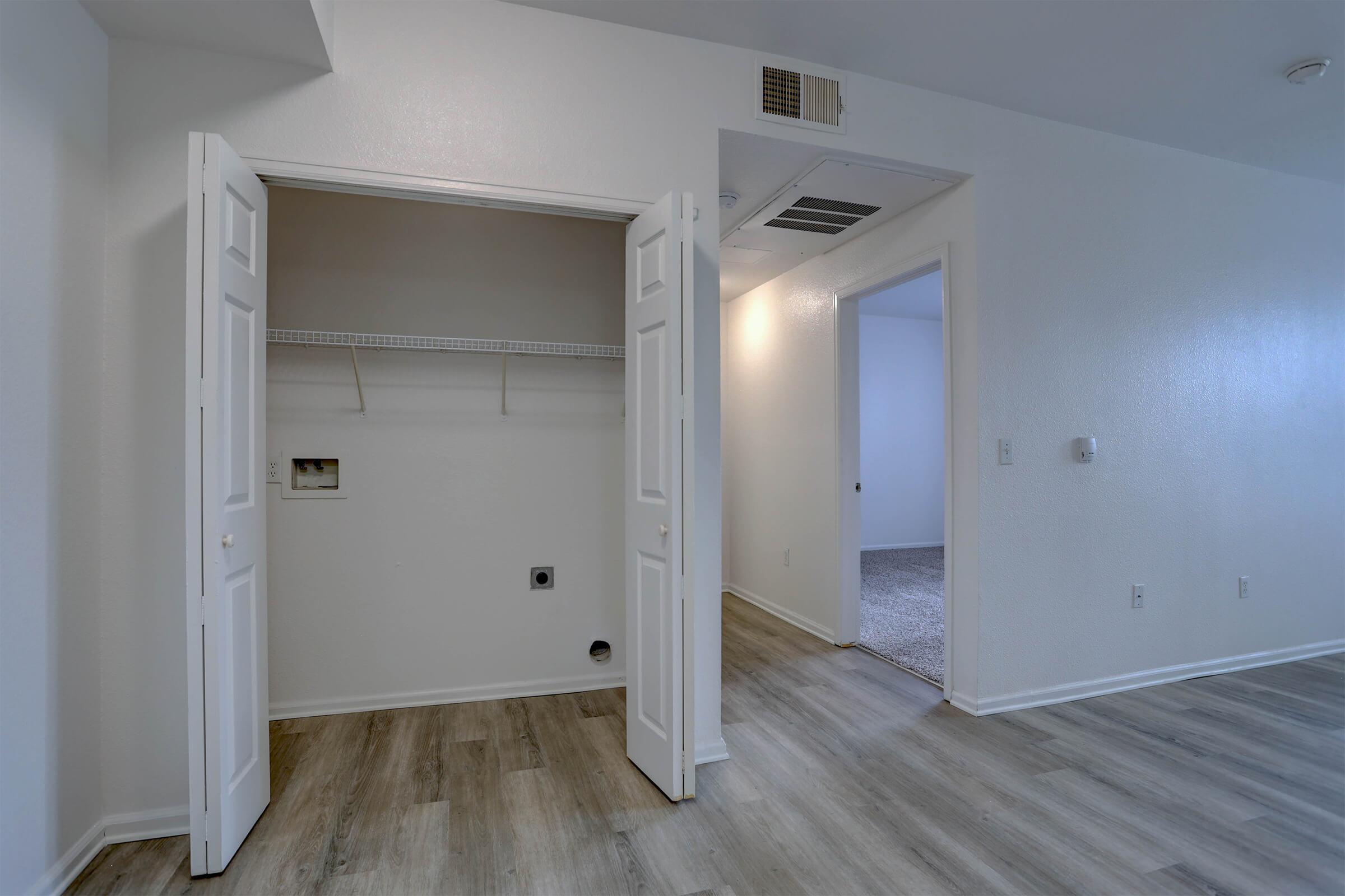 A spacious room featuring an open closet with double doors, light wood flooring, and white walls. A small utility area is located within the closet, and there is a doorway leading to another room in the background. Natural light brightens the space, creating a welcoming atmosphere.