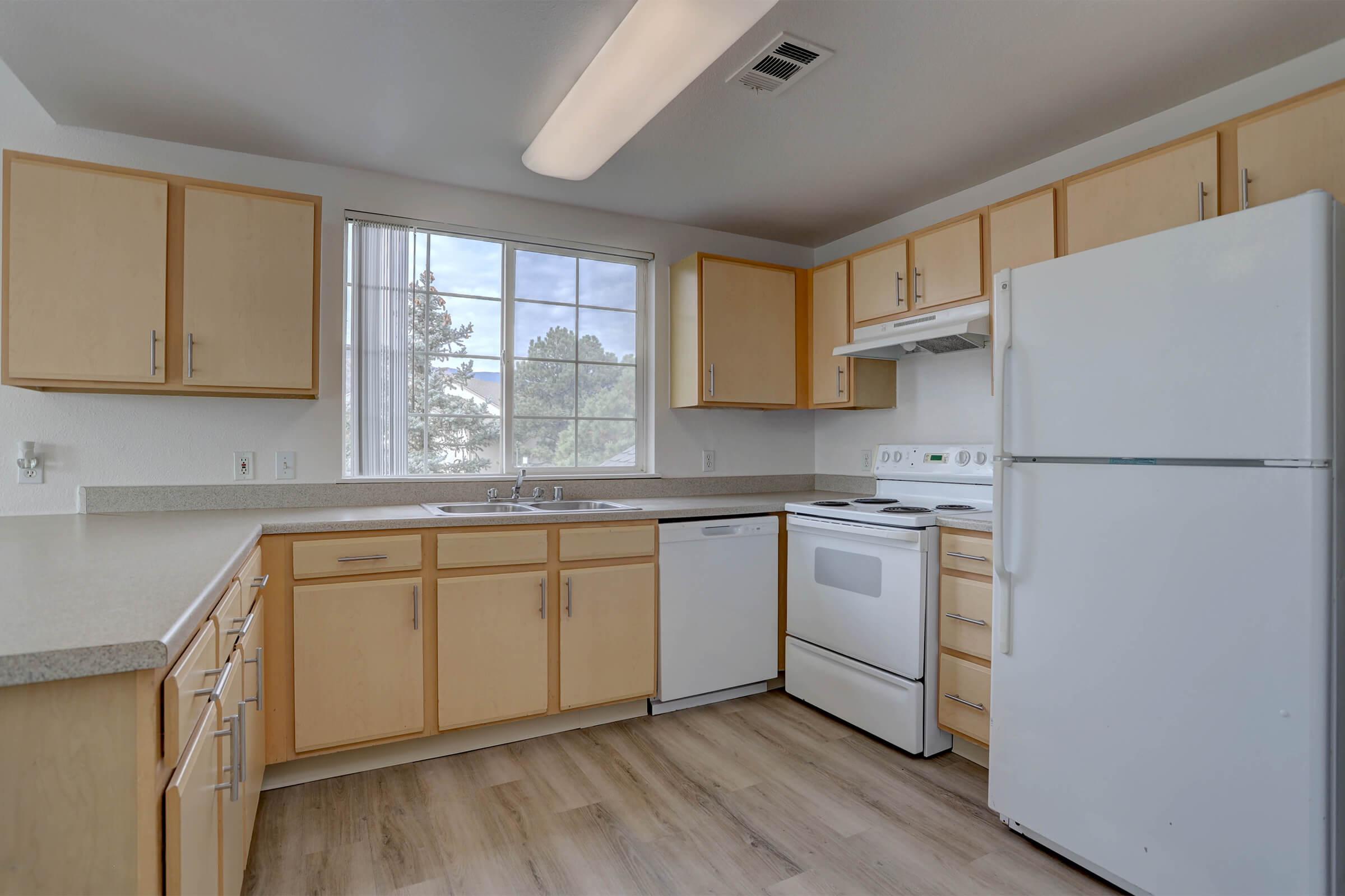 Bright kitchen with wooden cabinets, a large window allowing natural light, and modern appliances including a refrigerator, oven, and dishwasher. The countertops are light and there's a light-colored floor, creating a clean and airy atmosphere.