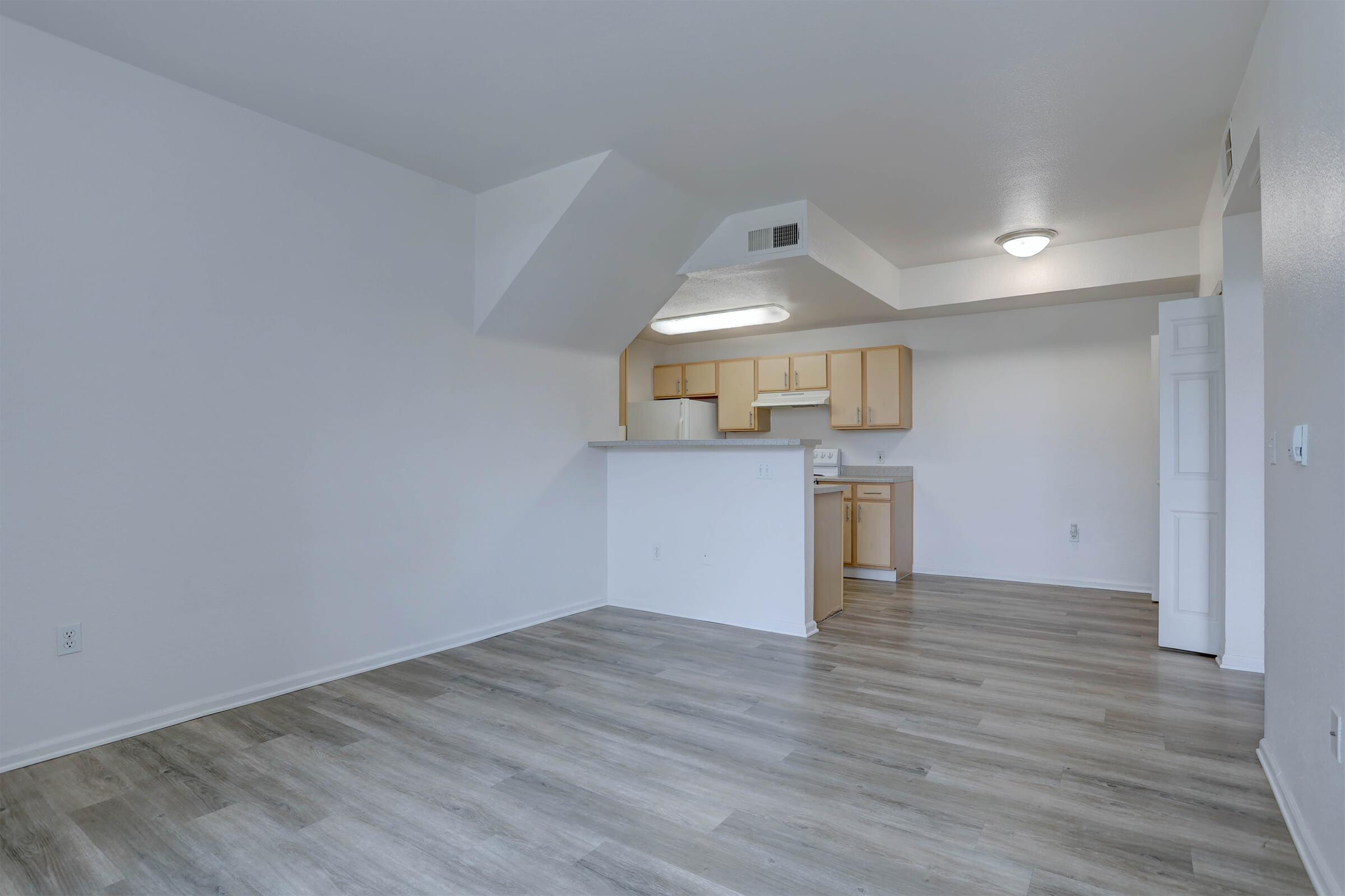 Spacious interior of an apartment featuring a light-colored wall, modern flooring, and a kitchen area with wooden cabinets. The room has ample natural light and an open layout, creating a welcoming atmosphere.