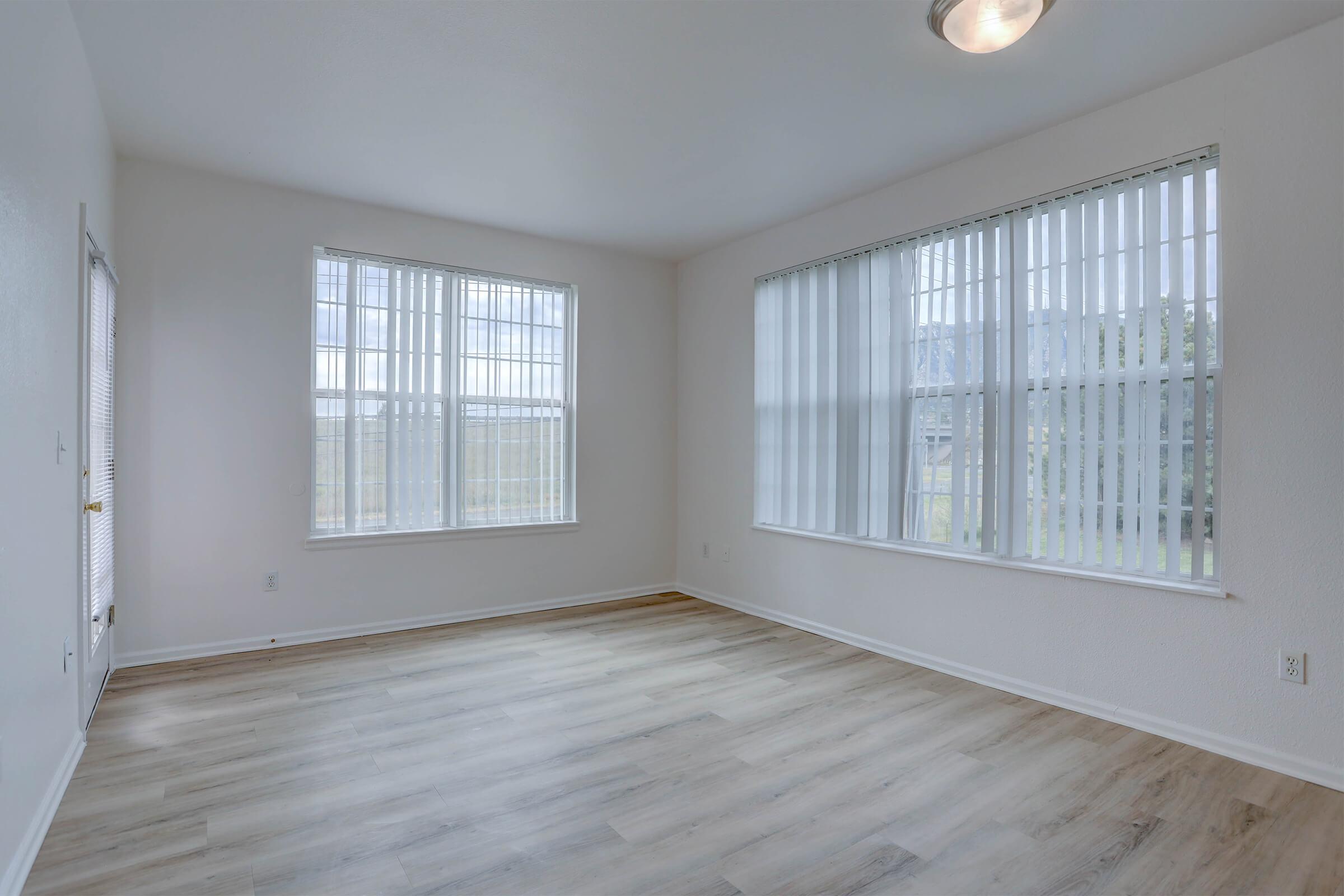 Bright, empty room with large windows featuring white vertical blinds. Light-colored wooden flooring complements the white walls. A modern ceiling light is visible, and there is a door on the left leading outside. The space is clean and spacious, ideal for various furniture arrangements.