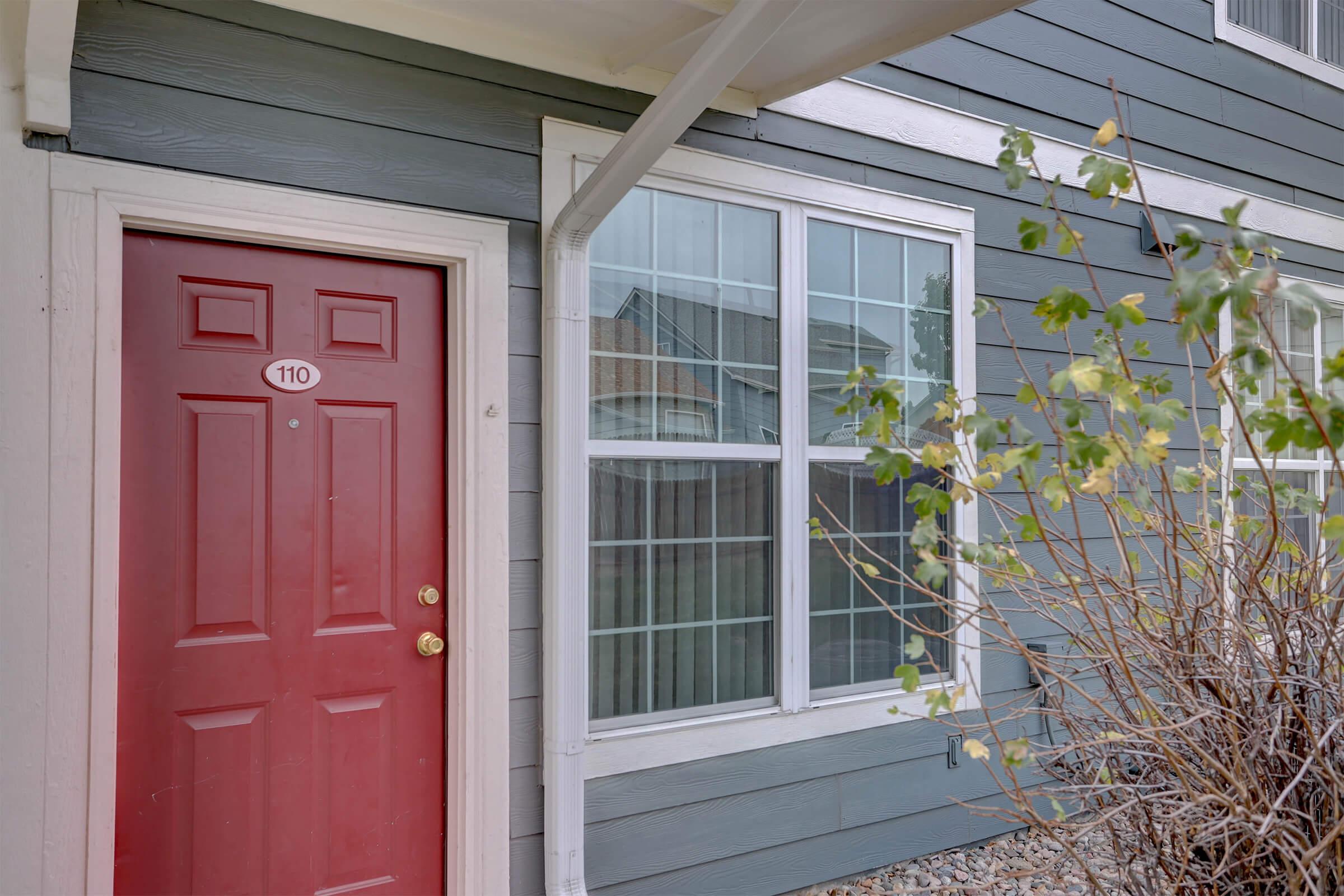 A close-up view of a red front door labeled "110" on a gray exterior wall. To the right, there is a large window with multiple panes, and some shrubbery in the foreground. The setting appears to be an entrance to an apartment or townhouse.