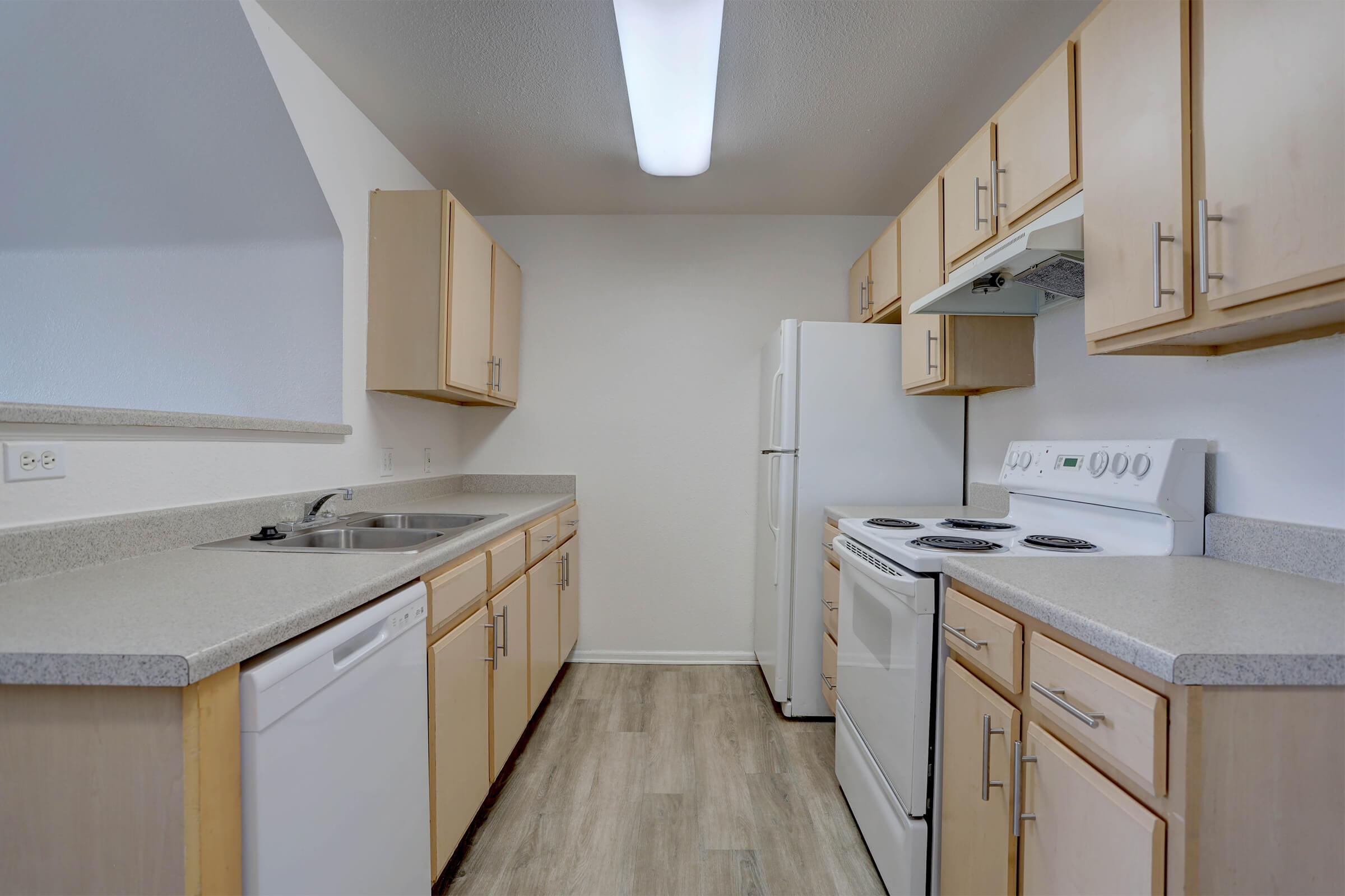 Modern kitchen with light wood cabinetry, gray countertops, and stainless steel appliances. Features a sink, dishwasher, oven, and refrigerator. Brightly lit with overhead lights, the space has a clean, minimalist design and a neutral color palette.
