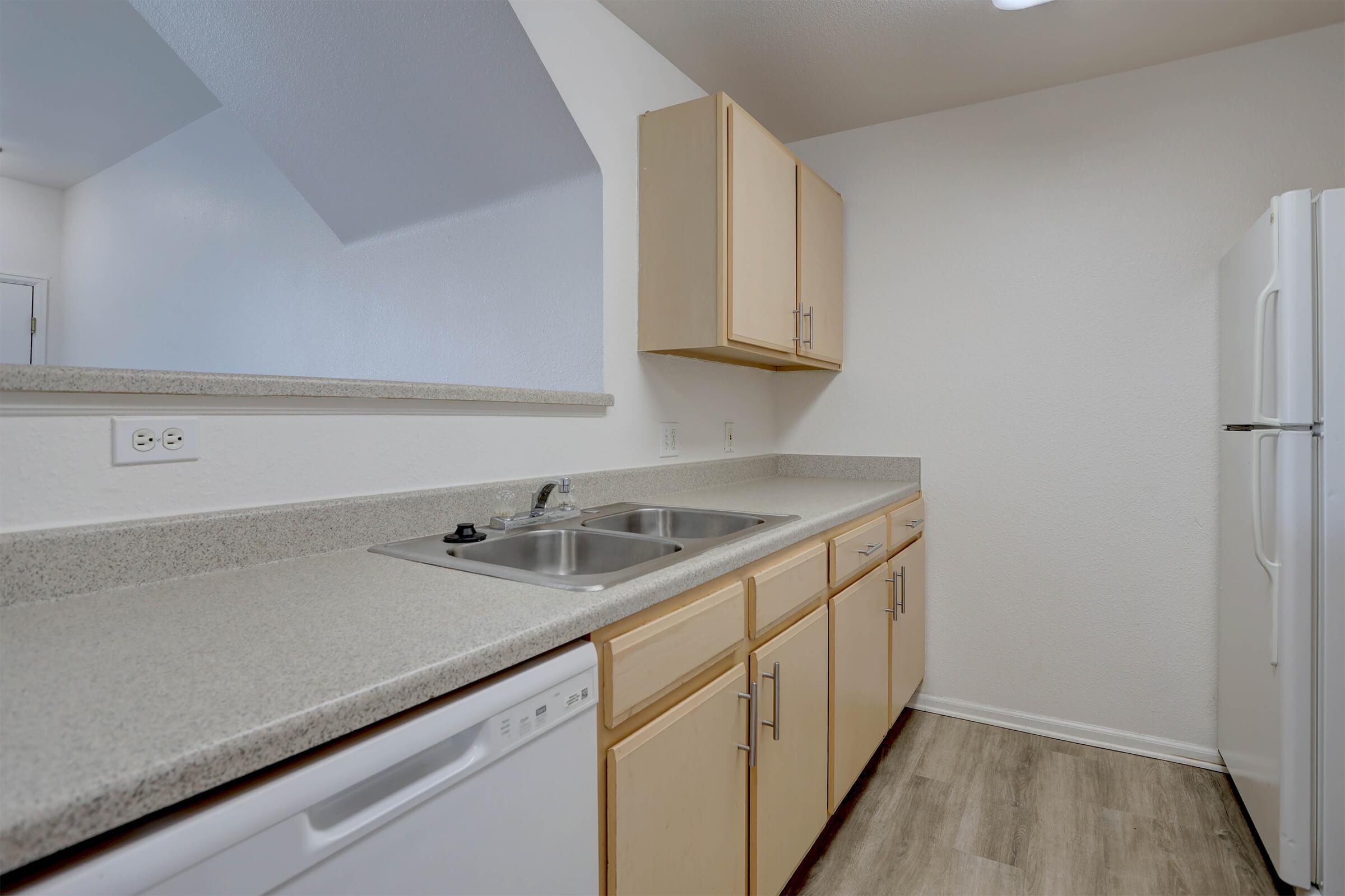 A modern kitchen featuring light-colored cabinets, a double sink, a dishwasher, and a white refrigerator. The countertop is a light gray material, and the flooring is a wood-like laminate. The walls are painted a neutral color, creating a bright and spacious atmosphere.