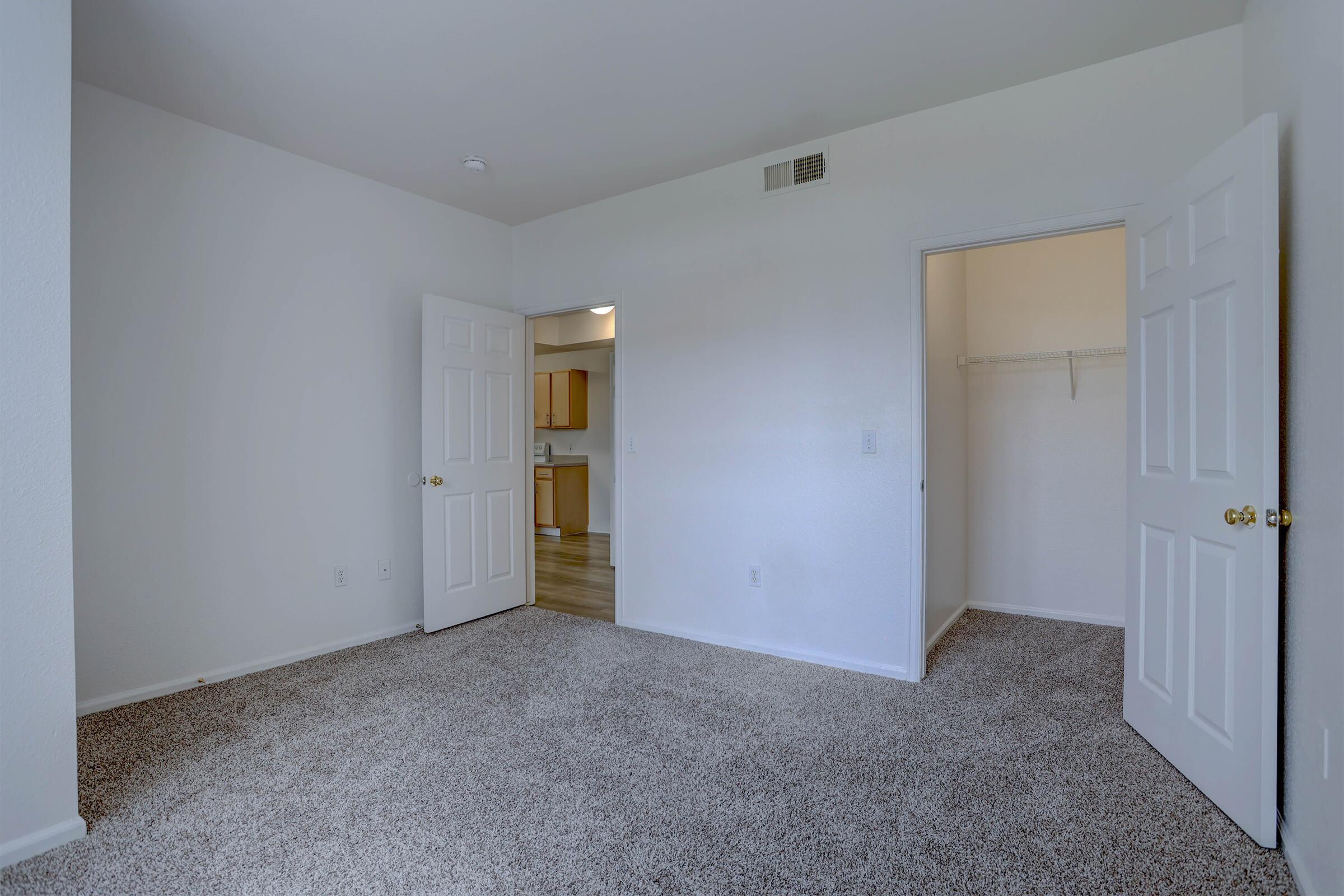 A spacious, empty room with light-colored walls and beige carpet. There are two open doors: one leading to a closet and another to a hallway with an adjacent room visible. The room is well-lit, featuring no furniture, creating a blank canvas for decoration.