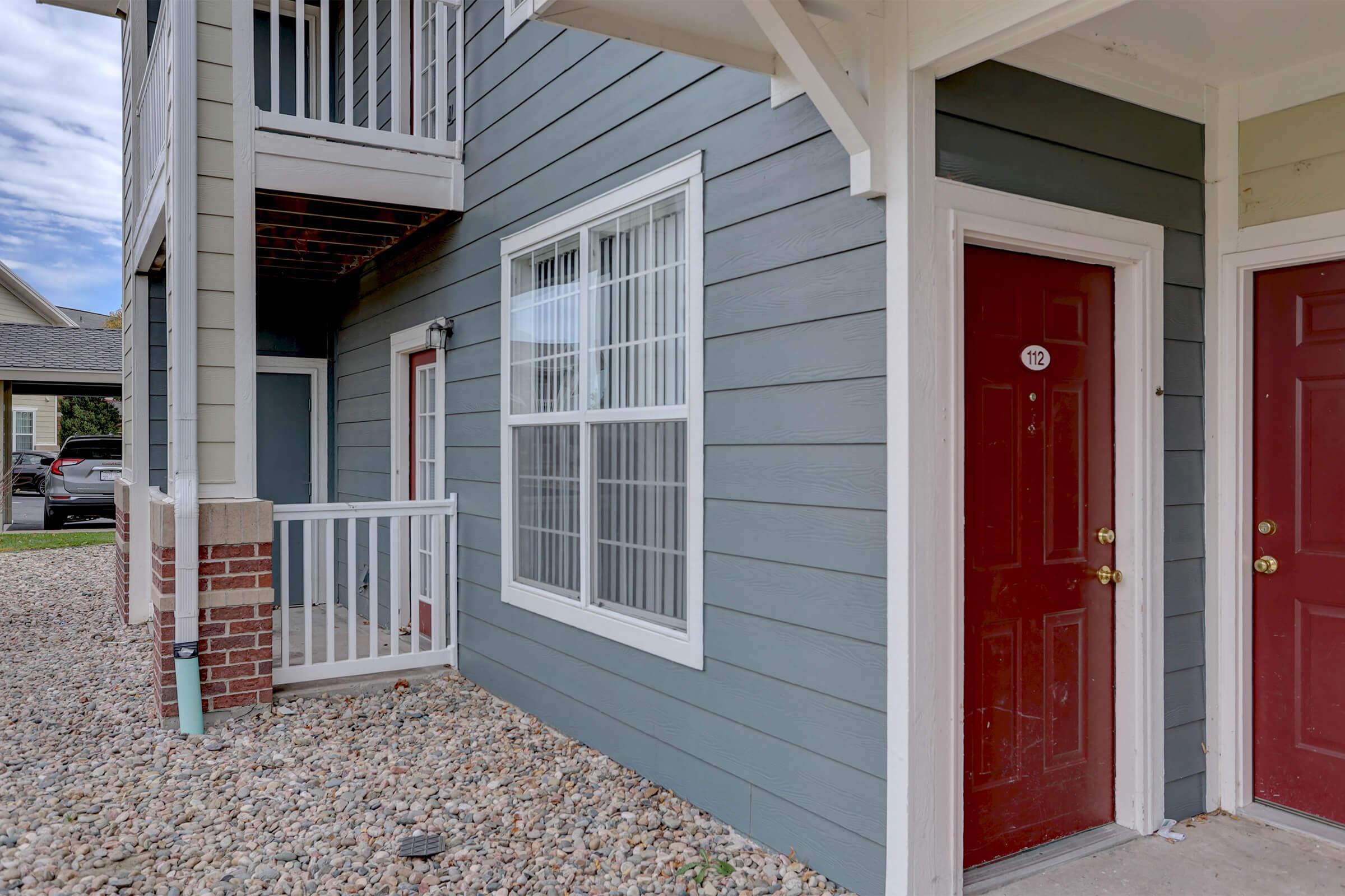 Exterior view of an apartment complex featuring a gray wooden siding, a ground-level entrance with a red door, and a small covered porch area with stairs leading to a second floor. Pebbles cover the ground in front of the building, and there are additional doors visible nearby.