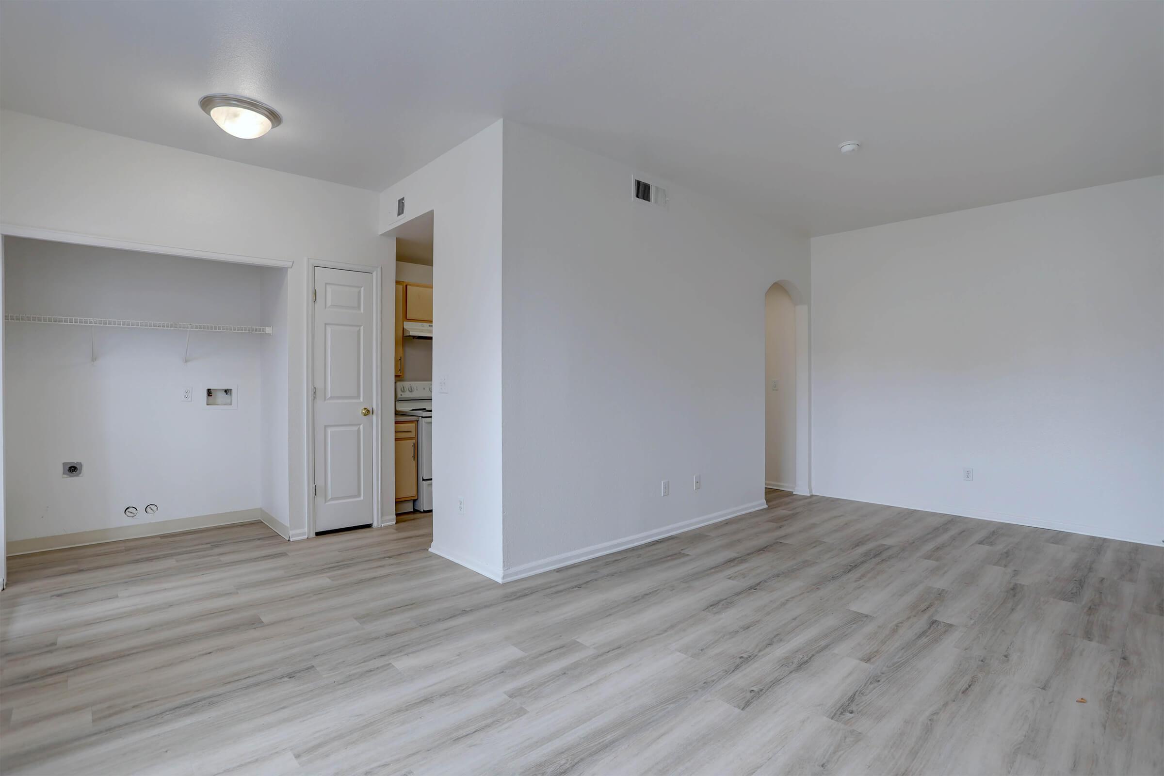 A bright, spacious living area featuring light-colored walls and new laminate flooring. The open layout connects to a small kitchen area, with a closet in the corner, creating a contemporary and inviting atmosphere. Natural light enhances the overall openness of the space.