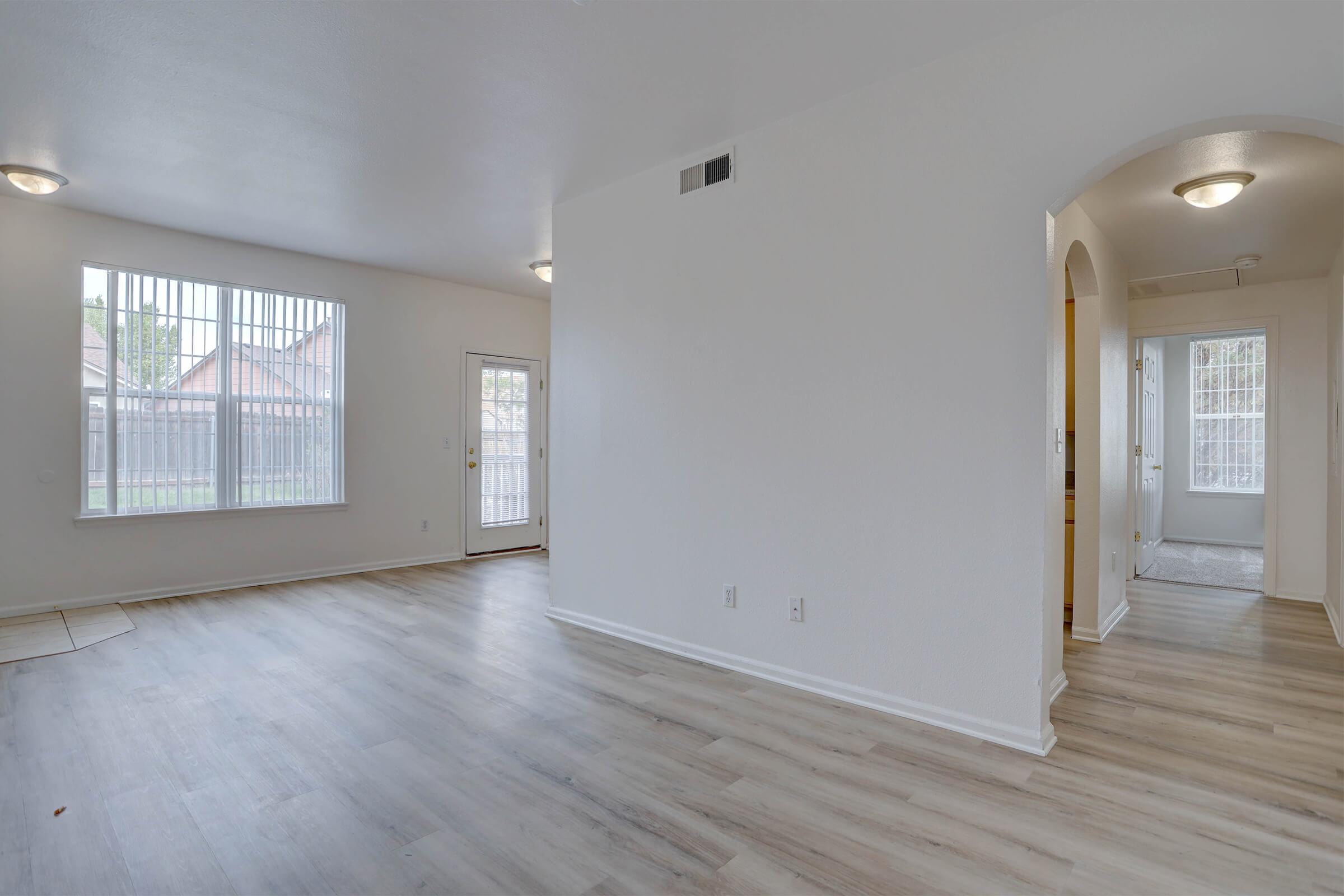 A spacious, empty room featuring light-colored walls and wooden flooring. There are large windows letting in natural light, a door leading outside, and an archway leading to another area. The space has a clean and modern feel, ideal for various uses.