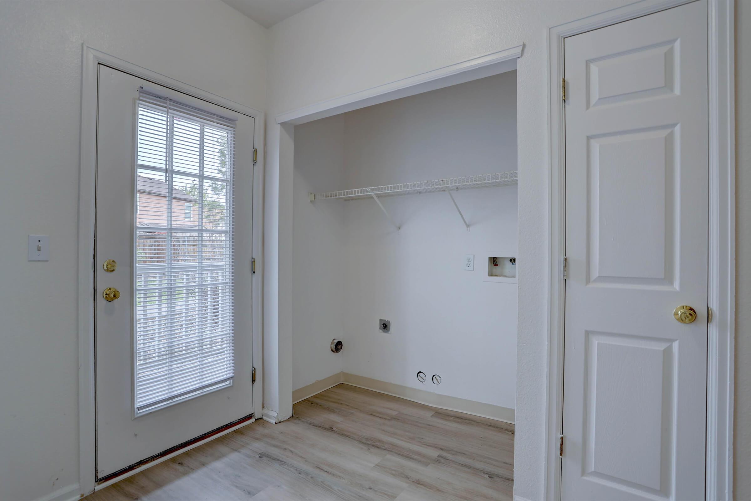 A small laundry room with light-colored walls, featuring a door with glass panels leading outside. It has a shelf mounted on the wall and is empty, with a floor of light laminate. Two closed doors are visible, one of which is a closet. Natural light enters from the door, making the space bright.
