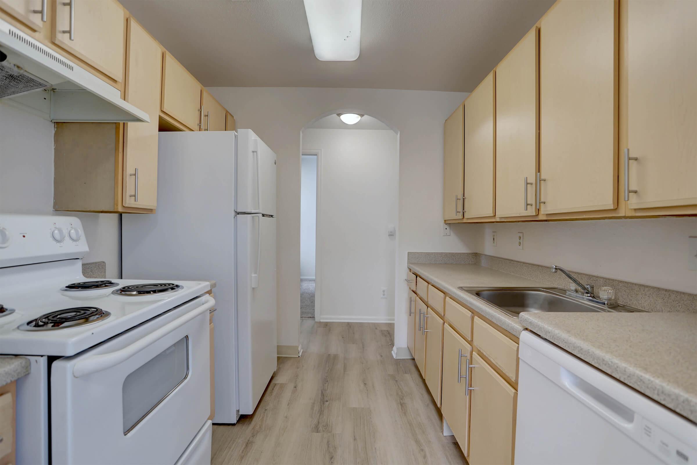 A view of a modern kitchen featuring light wood cabinetry, a white refrigerator, a stove with an oven, and a sink. The floor has a light wood finish, and there's an archway leading to another room. Natural light illuminates the space, creating a bright and inviting atmosphere.