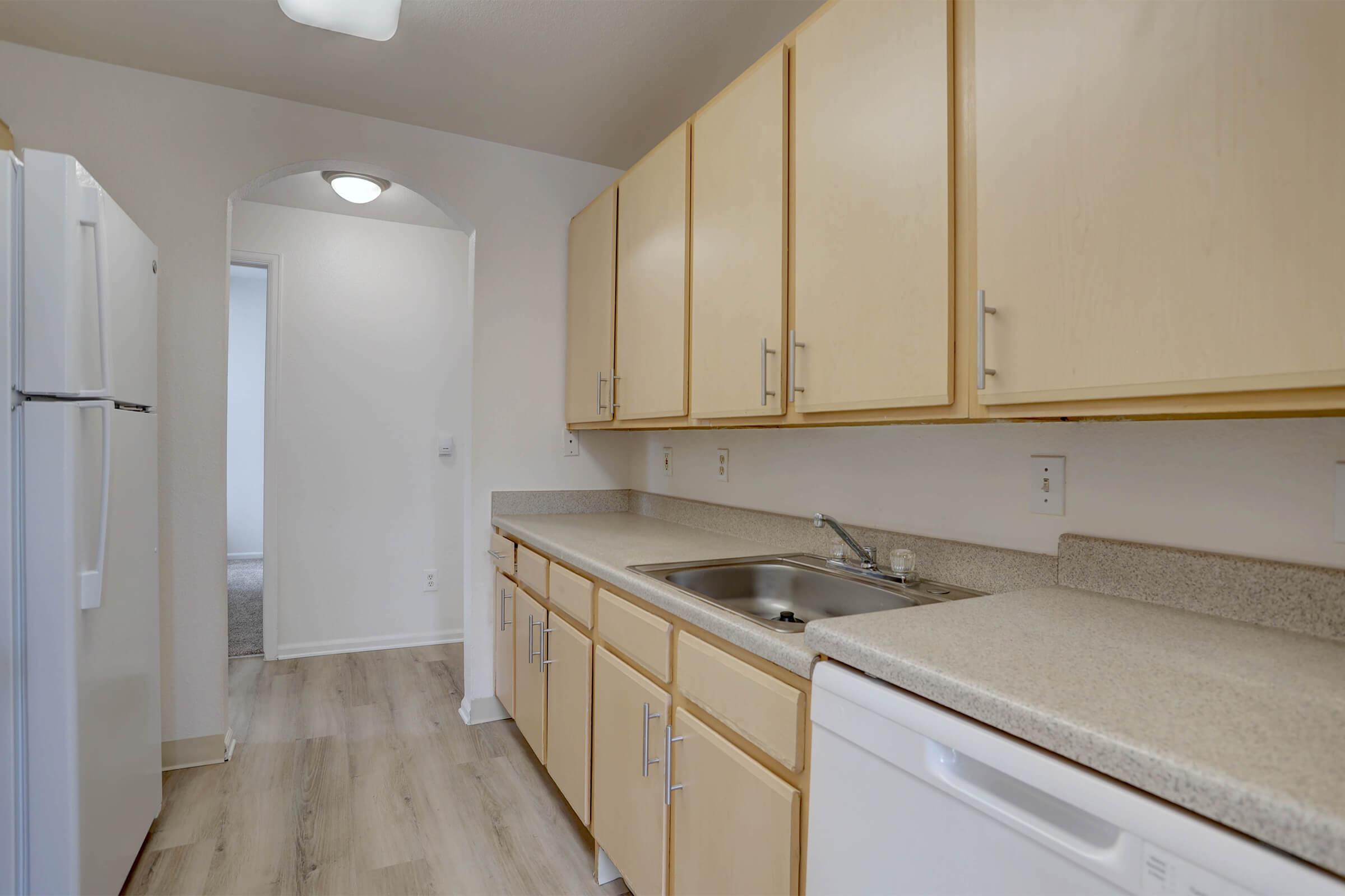 A modern kitchen featuring light wood cabinets, a stainless steel sink, and a countertop. The space includes white appliances, including a refrigerator and a dishwasher, with laminate flooring. A doorway leads to another room in the background, highlighting an open and functional layout.