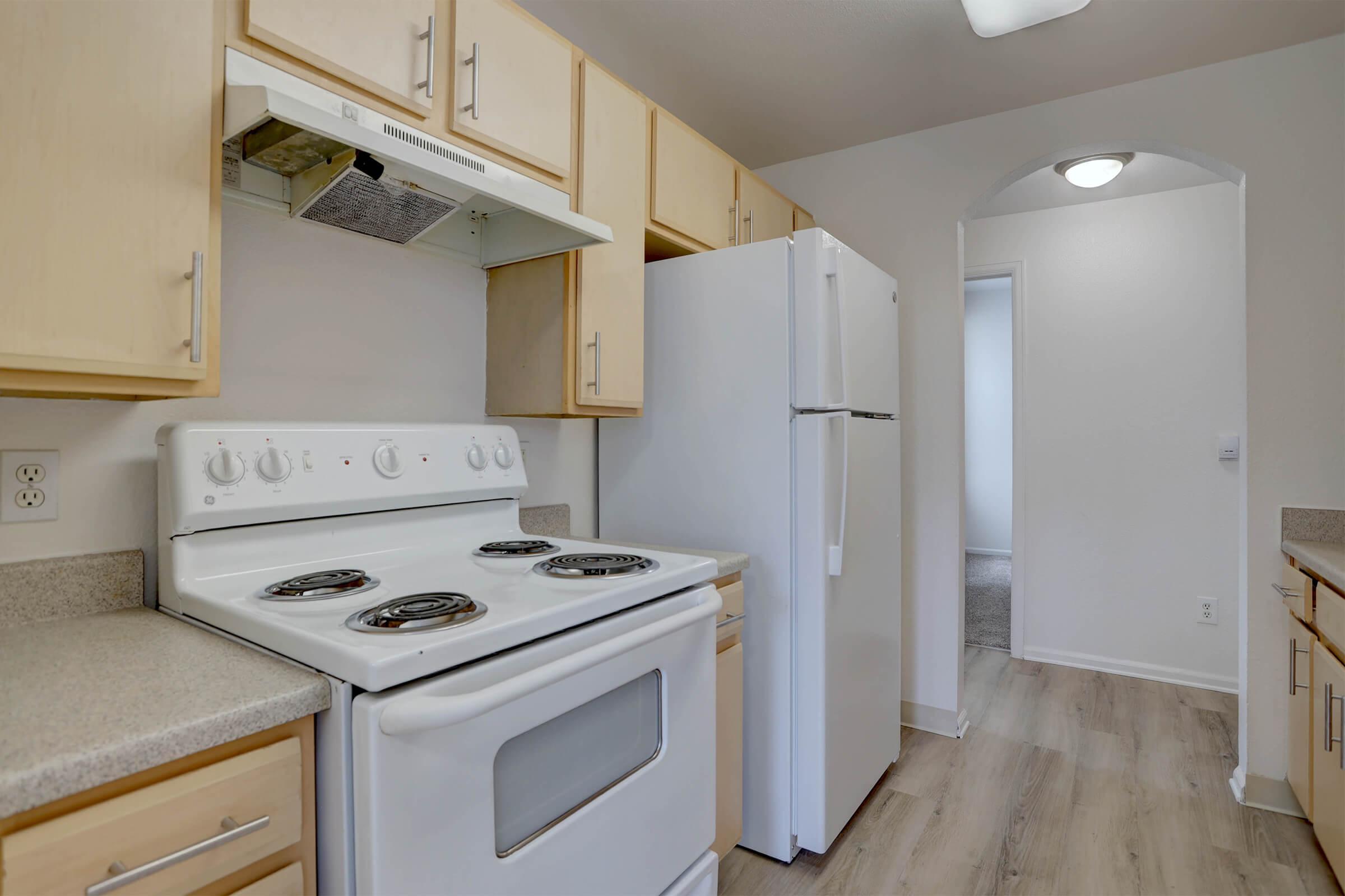 A modern kitchen featuring light wooden cabinets, a white stove, and a white refrigerator. The countertop is light gray, and the space has laminate flooring. A doorway leads to another room, and the kitchen is well-lit with natural light.
