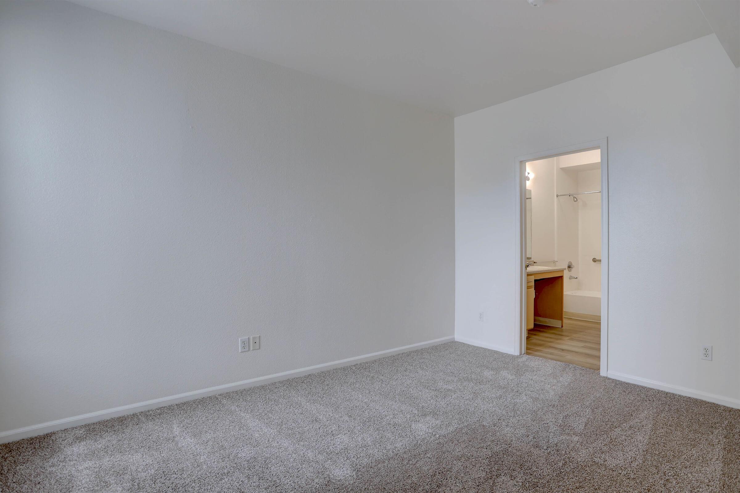 A spacious, empty room with light-colored walls and carpeted flooring. A doorway leads to a bathroom area in the background. The natural light entering from the doorway creates a bright, inviting atmosphere. The overall feel is clean and ready for personalization.