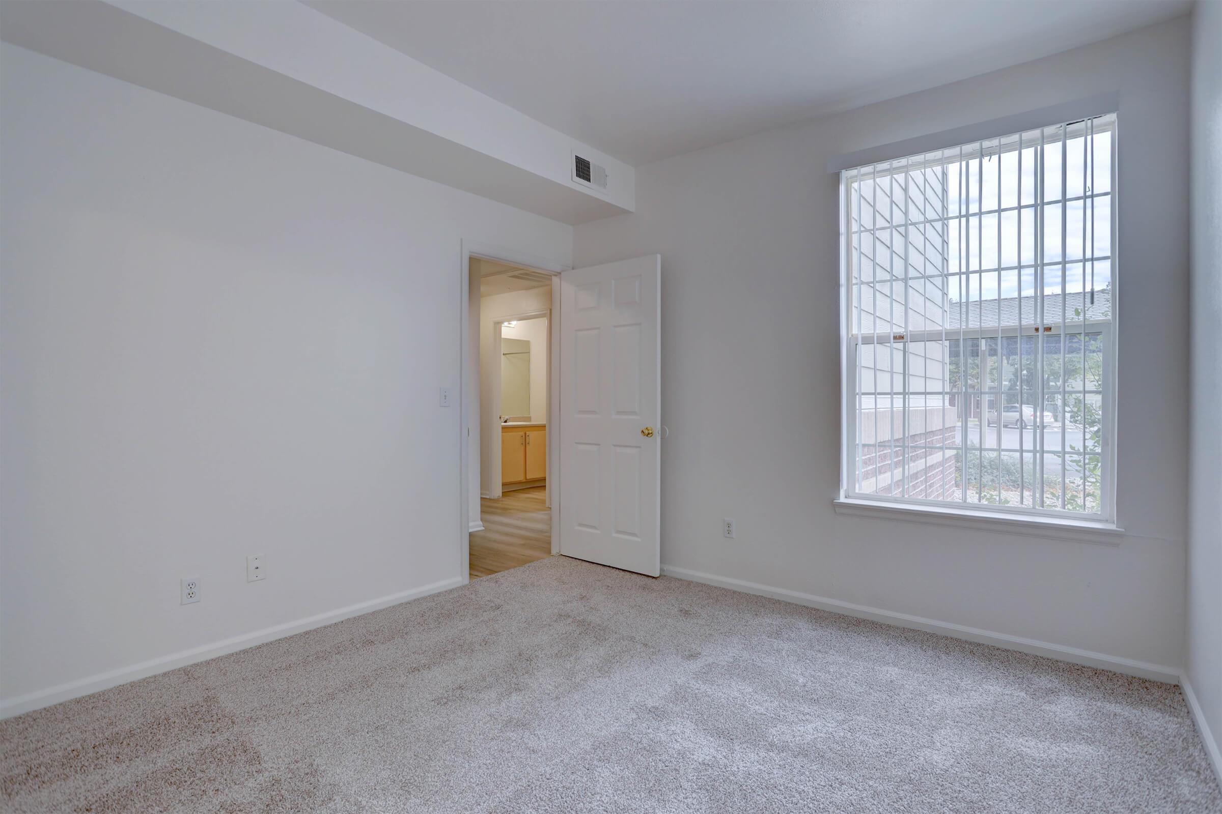 Empty room with beige carpet and white walls. A door on the left leads to another space, while a large window with blinds allows natural light to enter. The scene is minimalistic, creating a feeling of openness and potential for personalization.