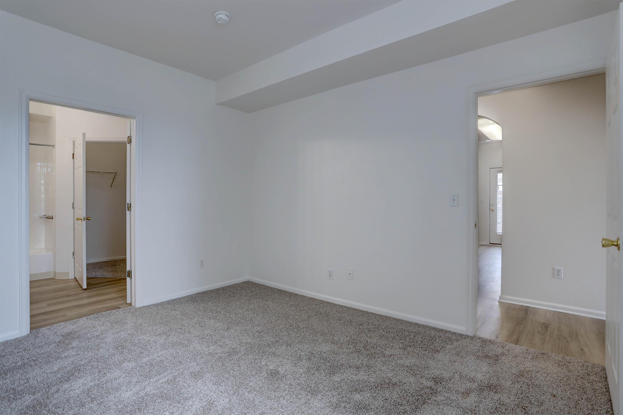 A spacious, empty room with gray carpet and white walls. There are two doorways leading to adjacent areas, one showing a bathroom with a door slightly open. Natural light is visible through the doorway on the right, suggesting another room beyond. The overall atmosphere is clean and modern.