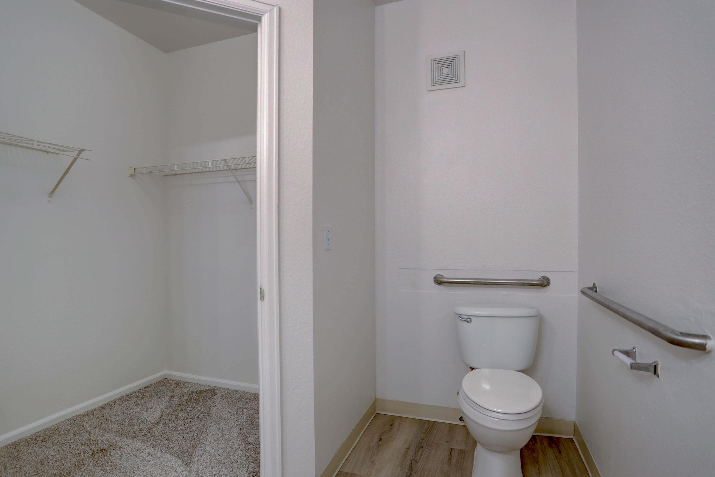A small bathroom featuring a toilet and a grab bar, with a closet space adjacent to it. The walls are painted white, and there is light-colored flooring. The closet has white wire shelving and bare carpeted flooring, creating a clean and simple design.
