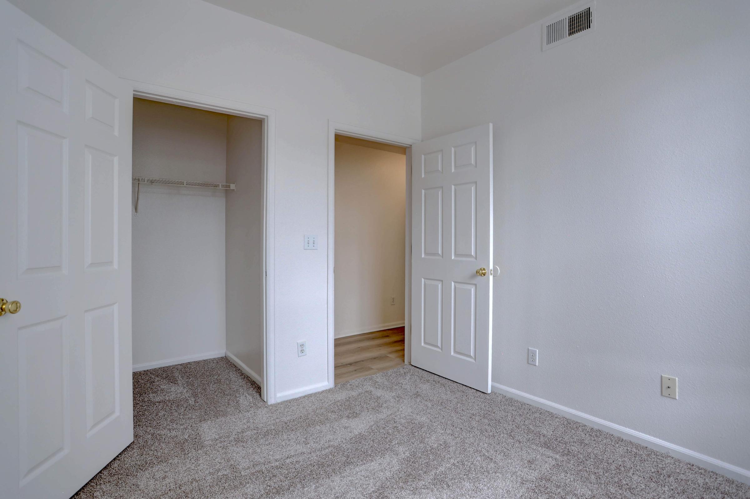A clean, empty bedroom with light-colored walls and carpeted floors. There are two doors, one leading to a closet and the other to a hallway, providing a spacious and bright atmosphere. The room is well-lit and minimalistic, suitable for various decorating styles.
