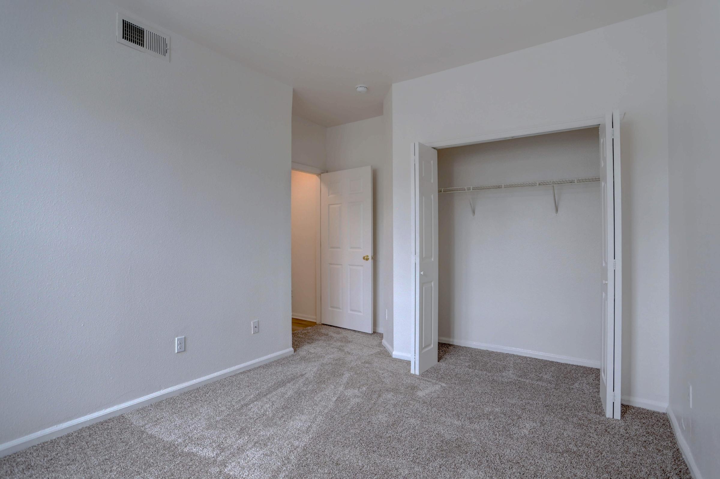 A small, empty room with beige carpet and white walls. There is a closet with a hanging rod and no doors, and an open door leading to another room. Natural light is coming in through an unseen window. The atmosphere is clean and simple, suitable for personal decoration.