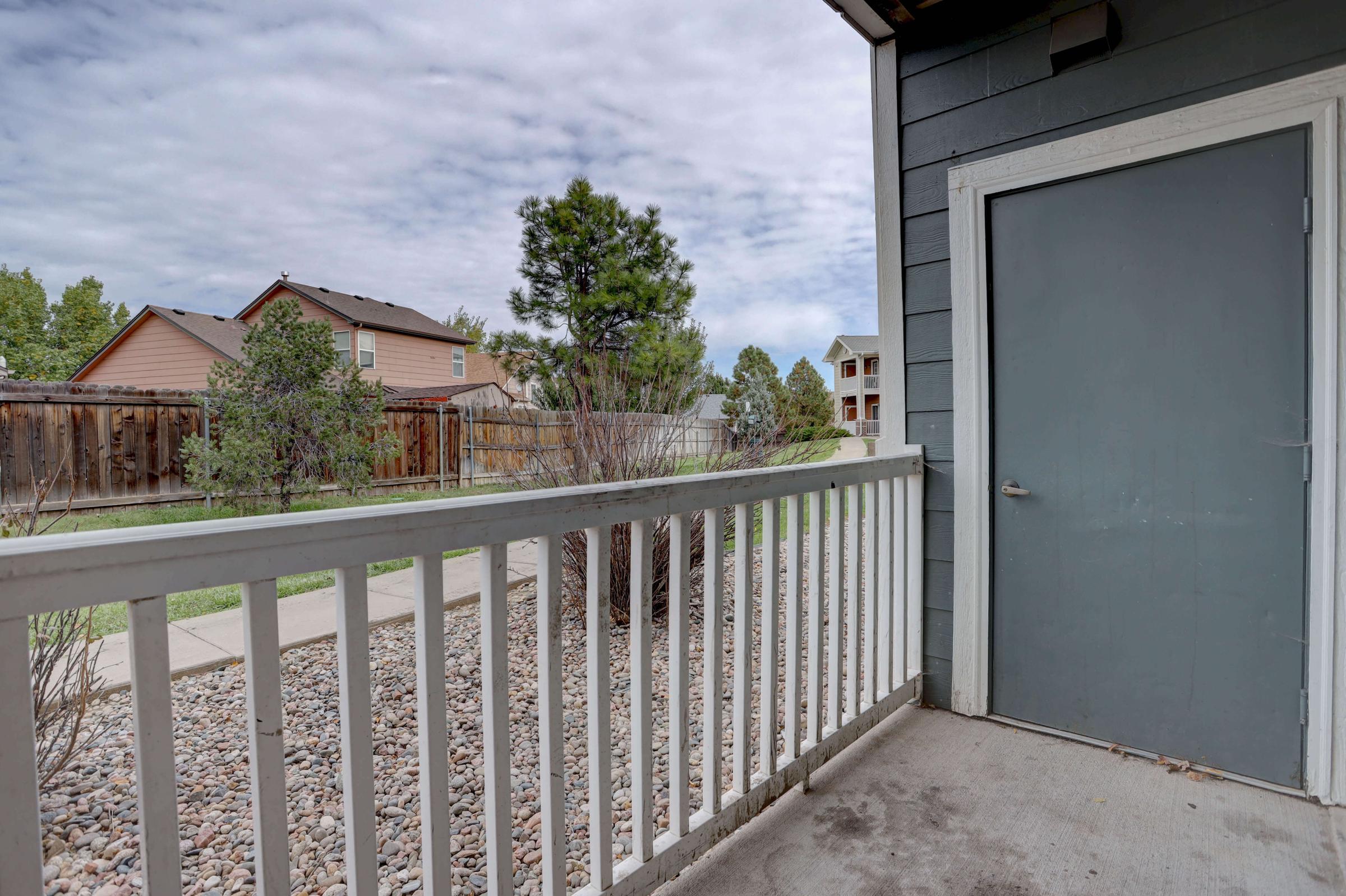 View from a porch featuring a gray door, railing, and landscaped area with small rocks. In the background, there are residential houses and trees under a partly cloudy sky.