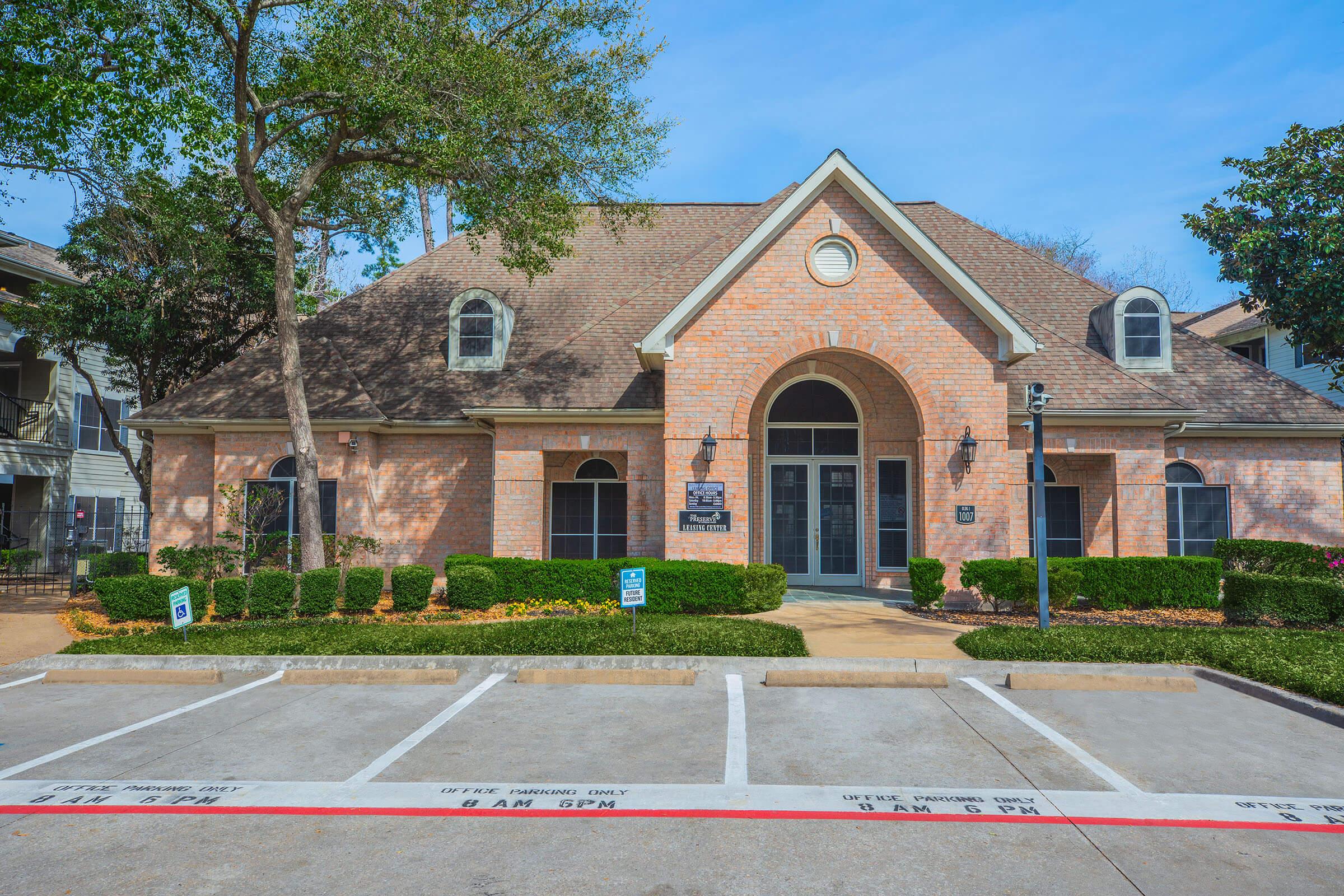 A brick building with a sloped roof and large arched entrance, surrounded by well-maintained landscaping. The front features several parking spaces with signs indicating parking regulations, and the sky is clear and blue.