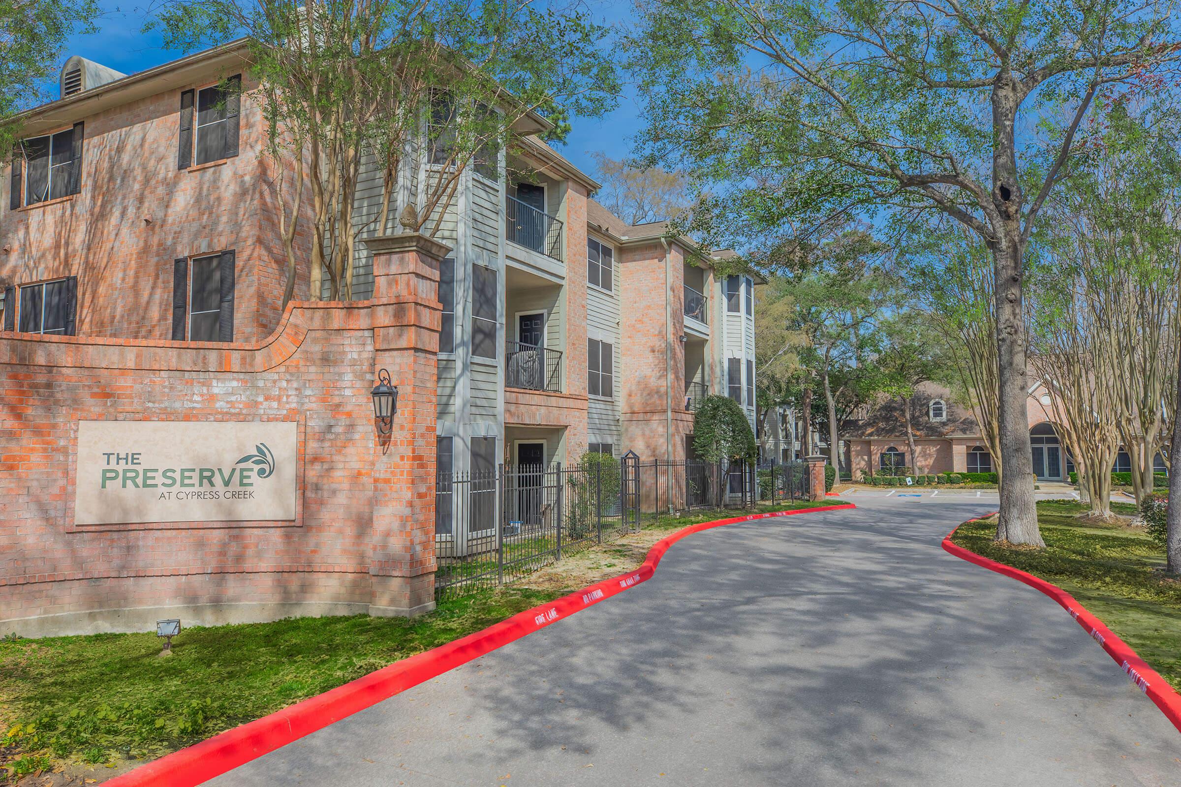 A well-maintained residential complex featuring brick and stucco buildings. The entrance displays a sign that reads "The Preserve at Cypress Creek." The driveway is edged with greenery and trees, leading into the property, which has a neatly landscaped surrounding area.