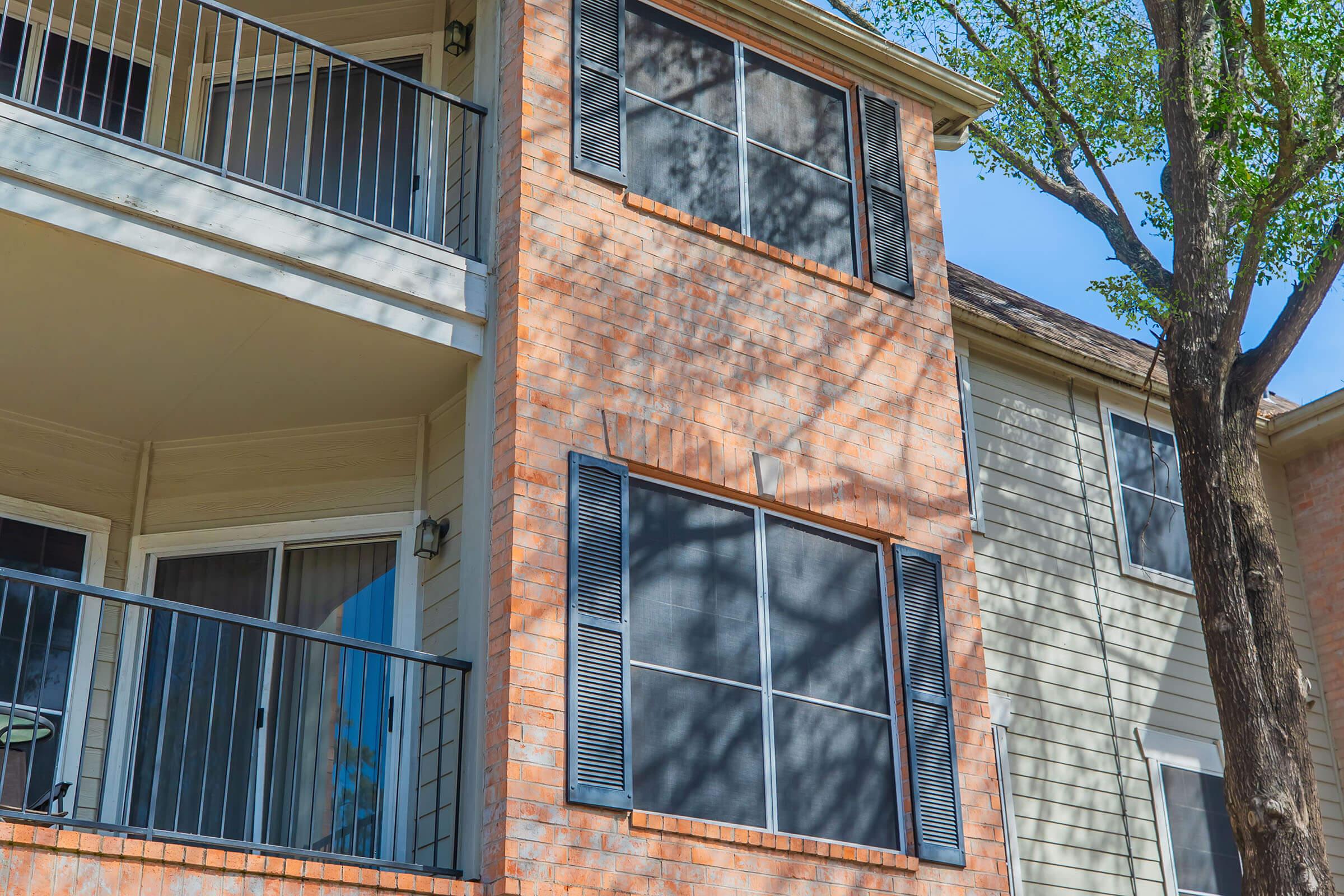 A close-up view of a two-story residential building with a brick and siding exterior. The building features several windows with black shutters and balconies, along with a tree casting shadows on the wall. The sky is clear and blue, suggesting a sunny day.