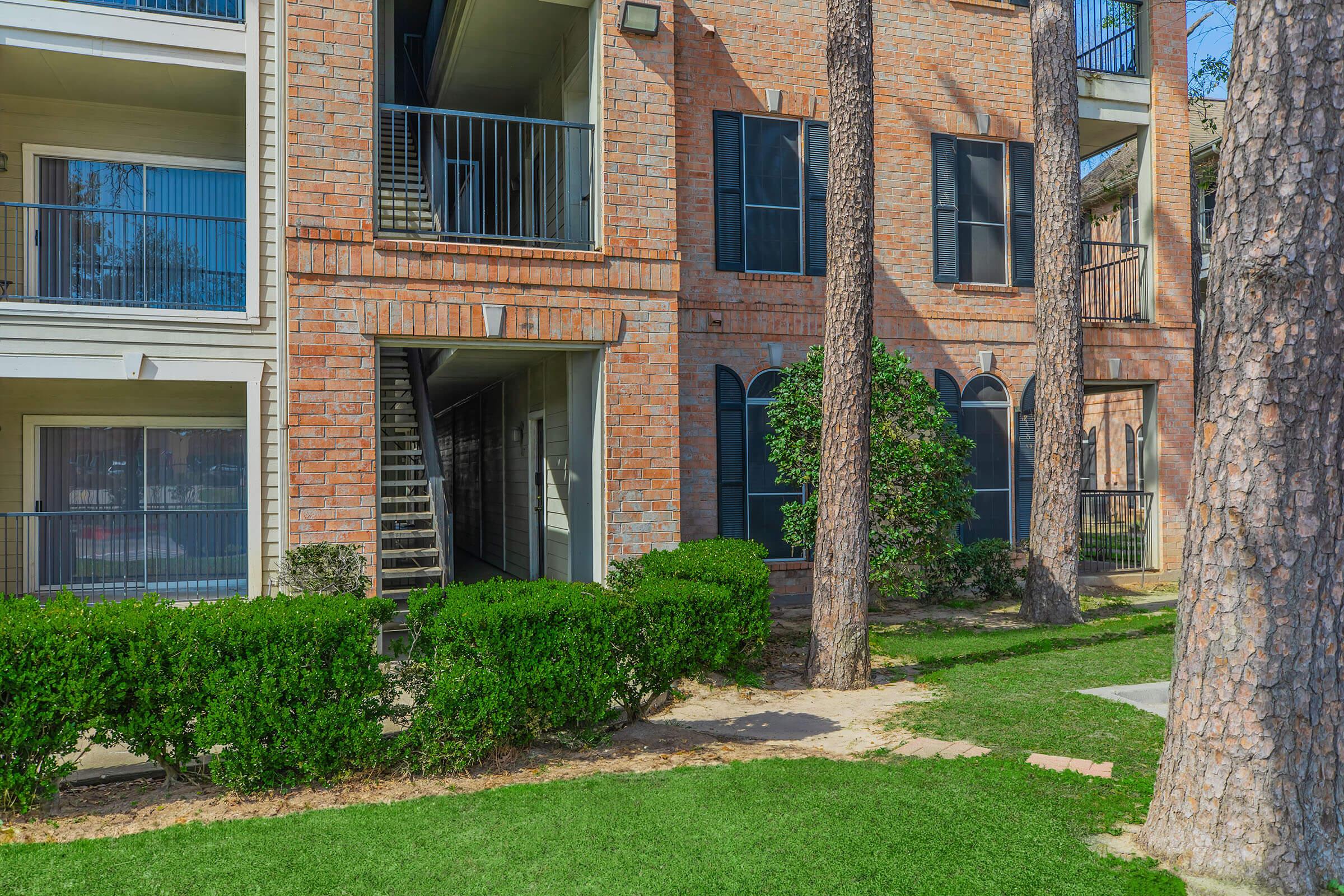 A residential building featuring brick exteriors, balcony railings, and large windows. The building is surrounded by well-maintained greenery, including bushes and trees. A pathway leads through the lawn, providing access to the entrance of the building.