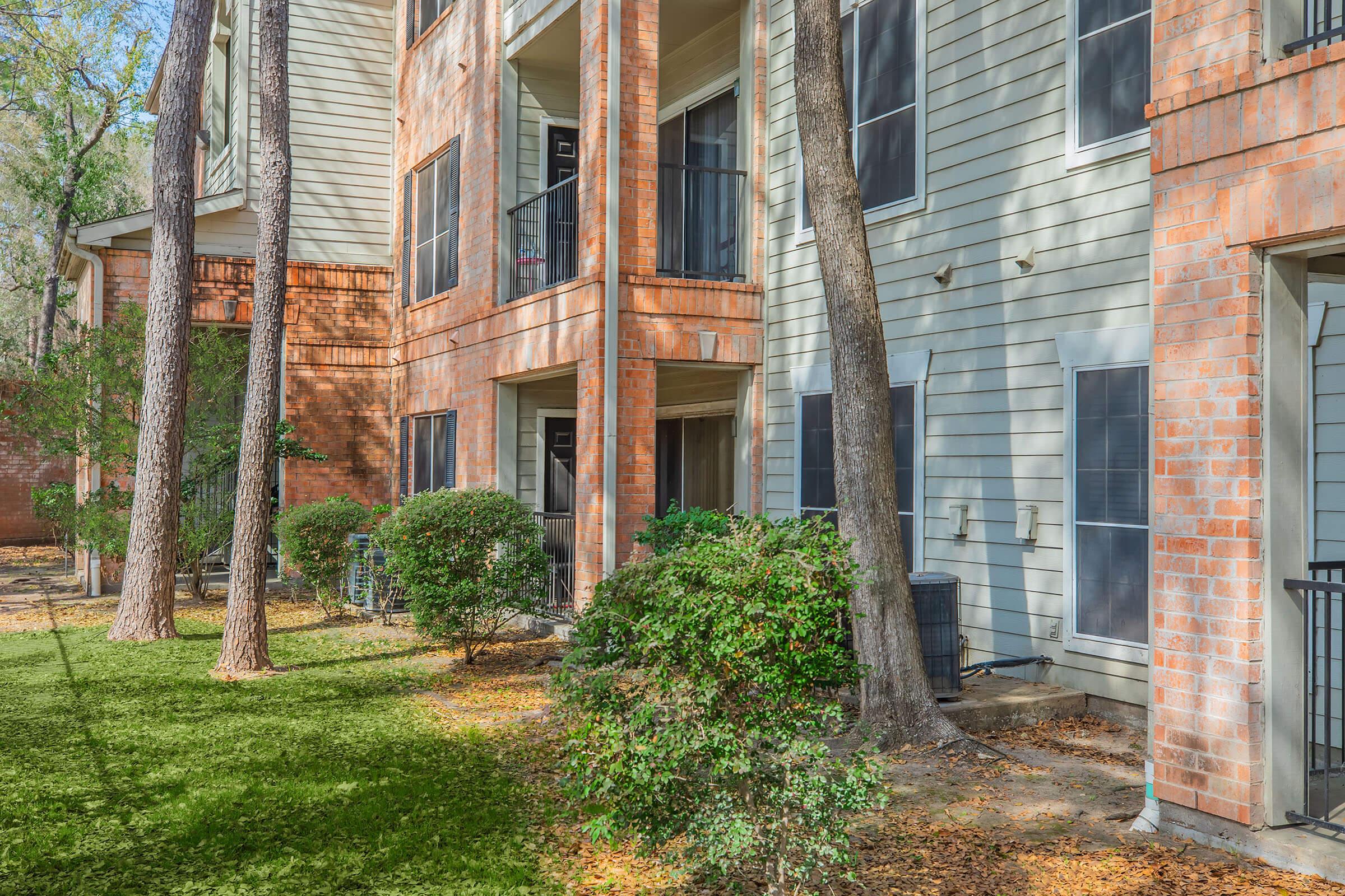 A view of an apartment complex featuring three-story buildings made of brick and siding. There are trees and bushes in the foreground, along with a green lawn scattered with fallen leaves. Balconies are visible on the buildings, and the setting appears to be tranquil and suburban.