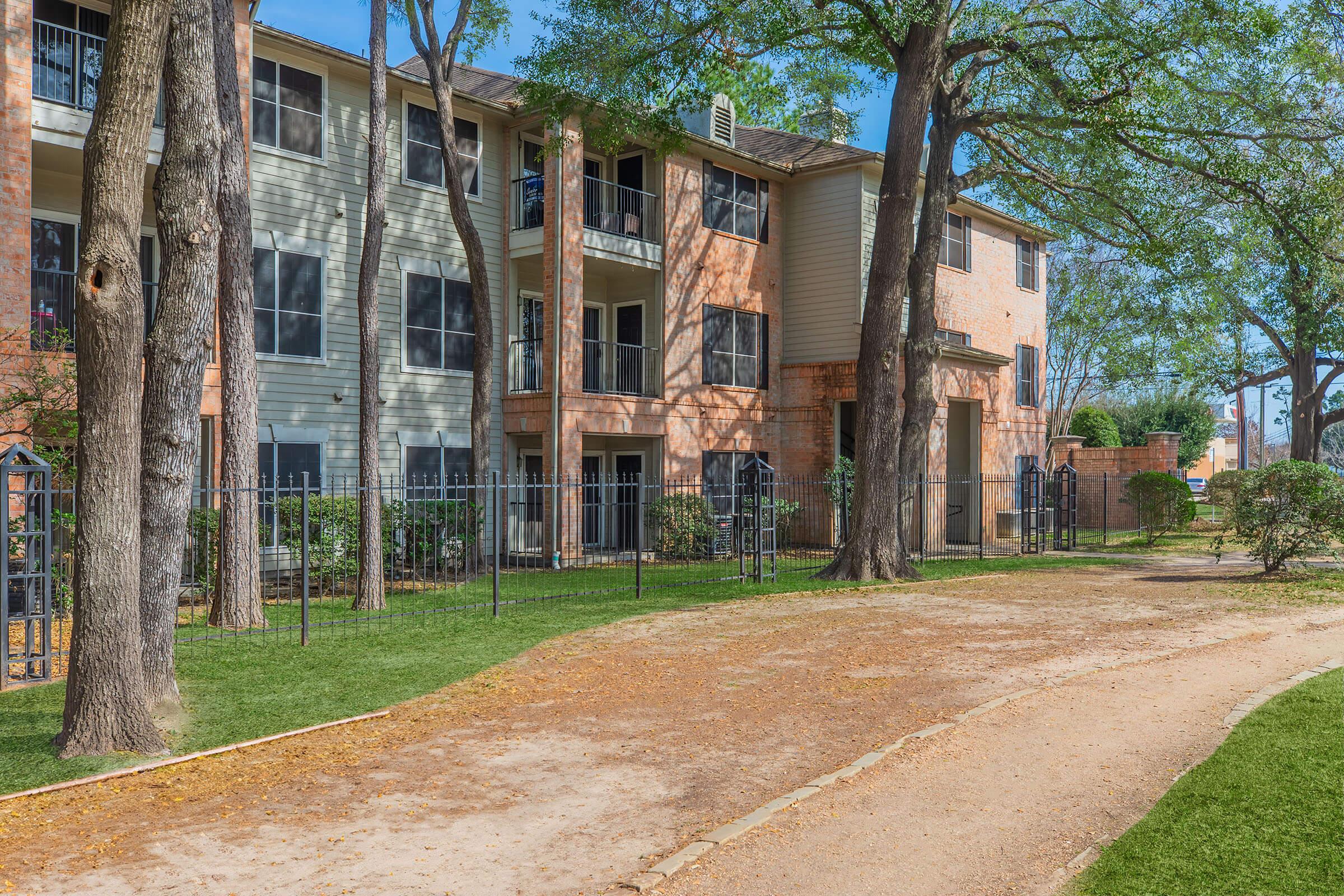 A view of a residential apartment complex surrounded by trees. The buildings feature balconies and a pathway lined with grass, leading through a well-maintained outdoor area. Clear blue skies add to the pleasant ambiance of the setting.