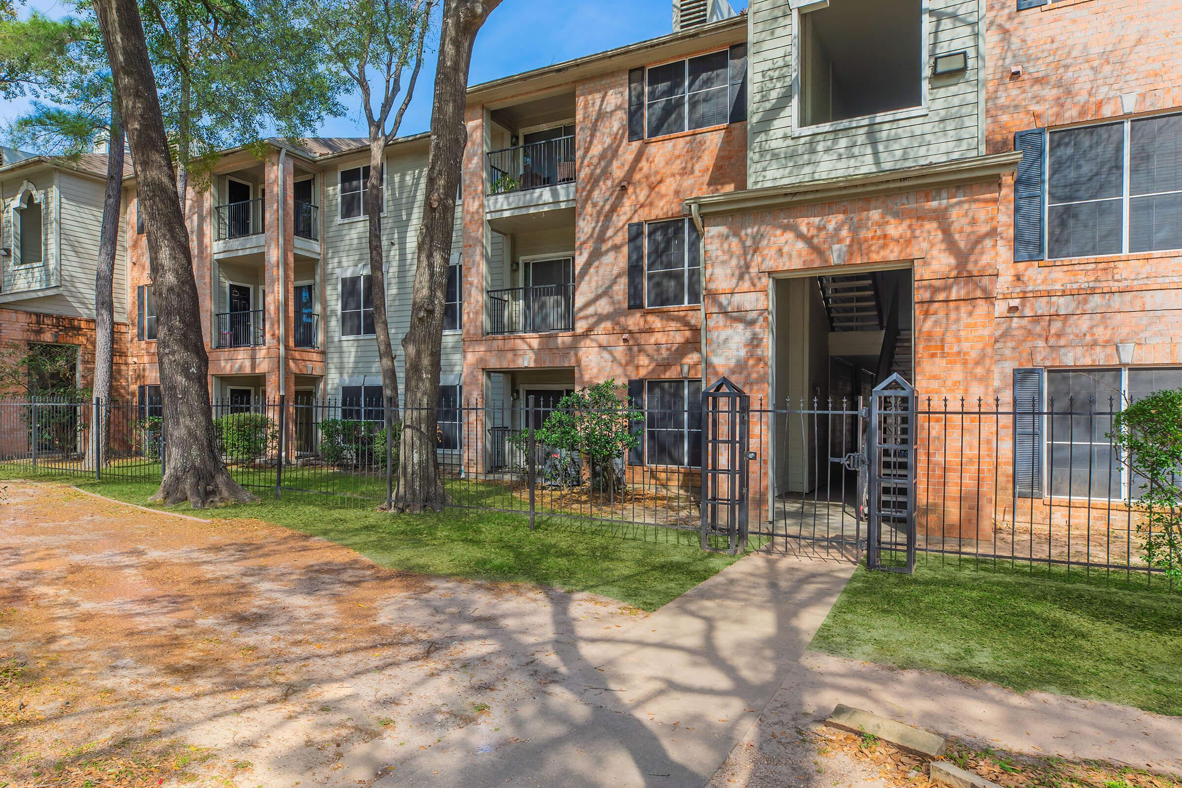 A view of an apartment complex featuring multiple stories with exterior brick and siding. The building is surrounded by trees and a gated entryway, with a pathway leading to the entrance. Sunlight filters through the foliage, casting shadows on the ground.