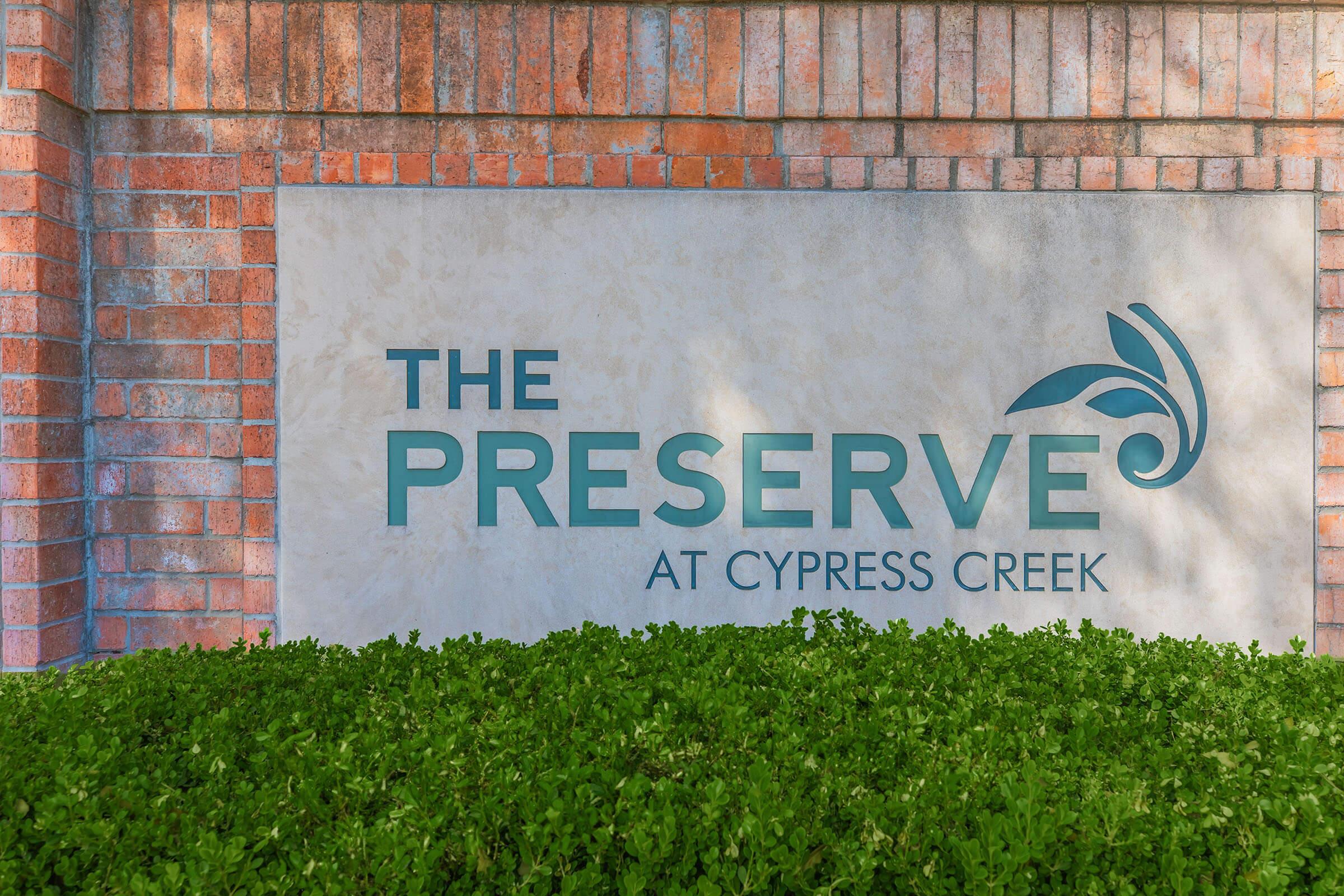 Sign for "The Preserve at Cypress Creek" displayed on a wall made of bricks, surrounded by green foliage. The sign features modern lettering with a stylized leaf motif.