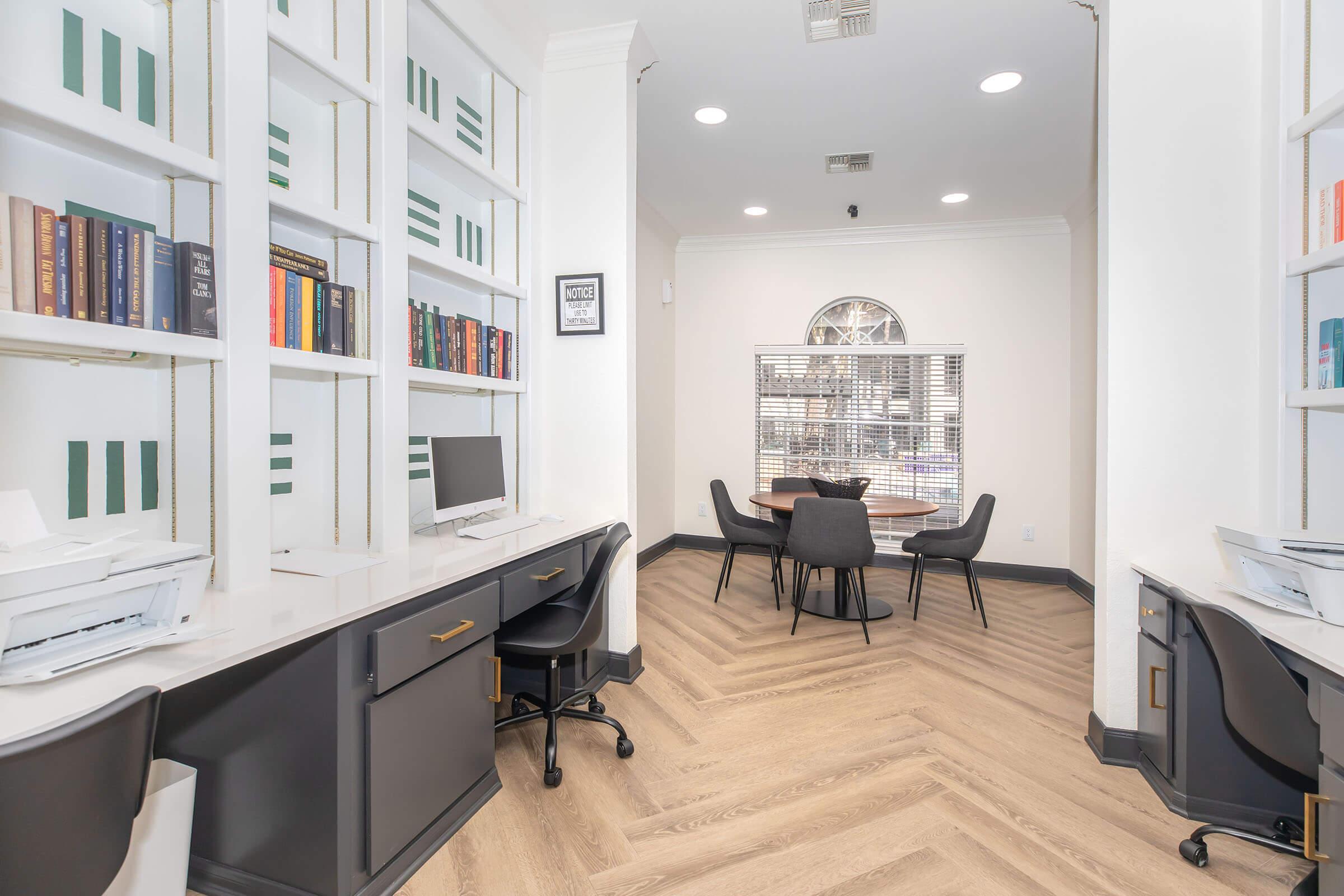 A modern office space featuring two desks with computers against white shelves filled with books. In the background, a round table with four chairs is visible, and large windows allow natural light to illuminate the room. The flooring is a stylish herringbone pattern, contributing to the contemporary aesthetic.