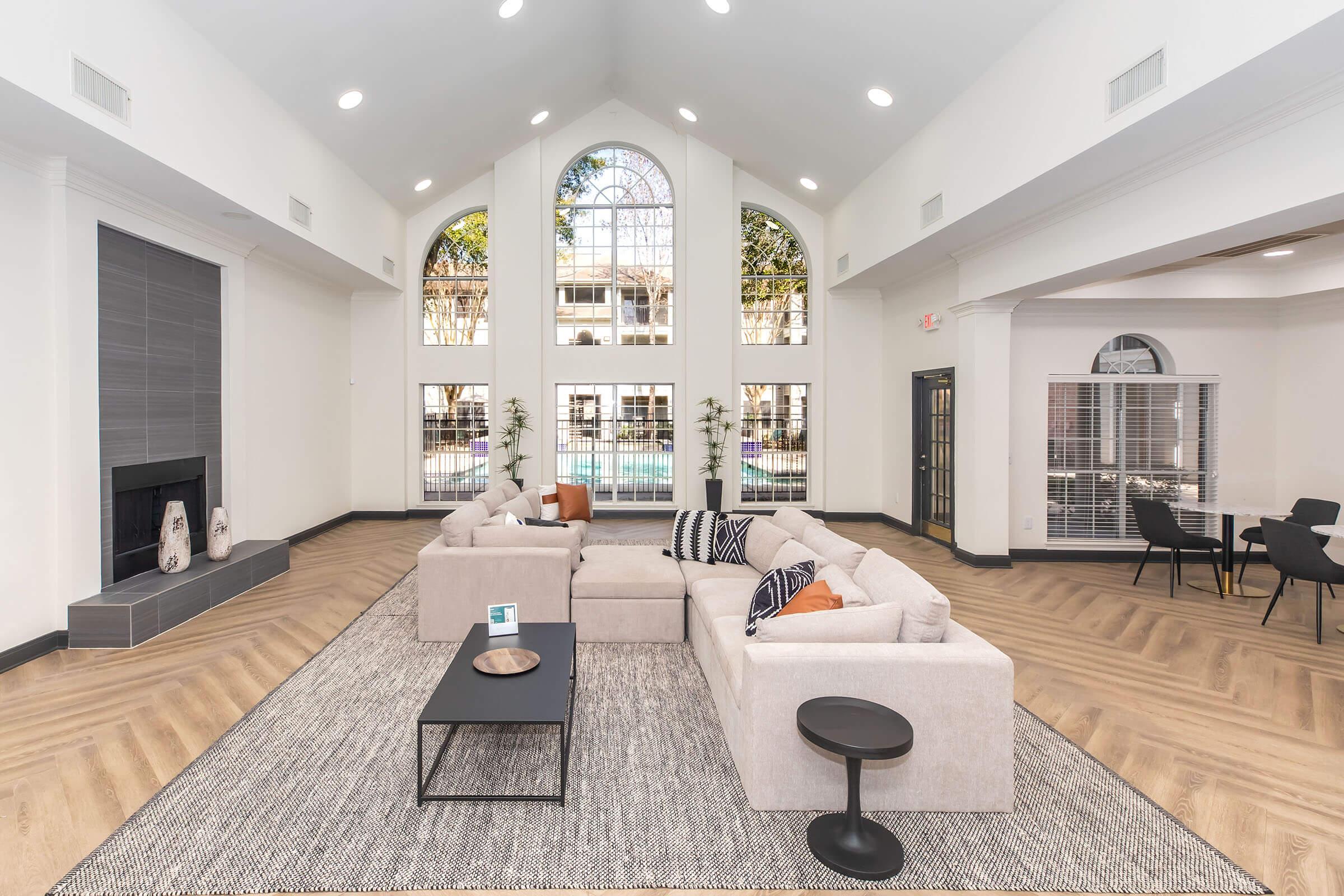 A spacious, modern living area featuring a light-colored sectional sofa, a coffee table, and a decorative bowl. Large windows allow natural light, showcasing a pool area outside. The walls are painted white, with a sleek fireplace on the left and a dining table set in the corner. Hardwood floors add warmth to the space.