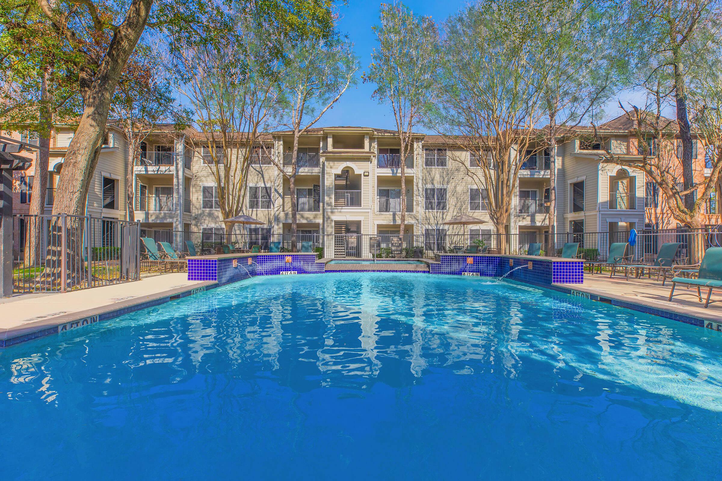 A clear swimming pool surrounded by lounge chairs and bordered by trees, with an apartment building in the background. The sun shines brightly, reflecting off the water's surface, creating a tranquil and inviting atmosphere.