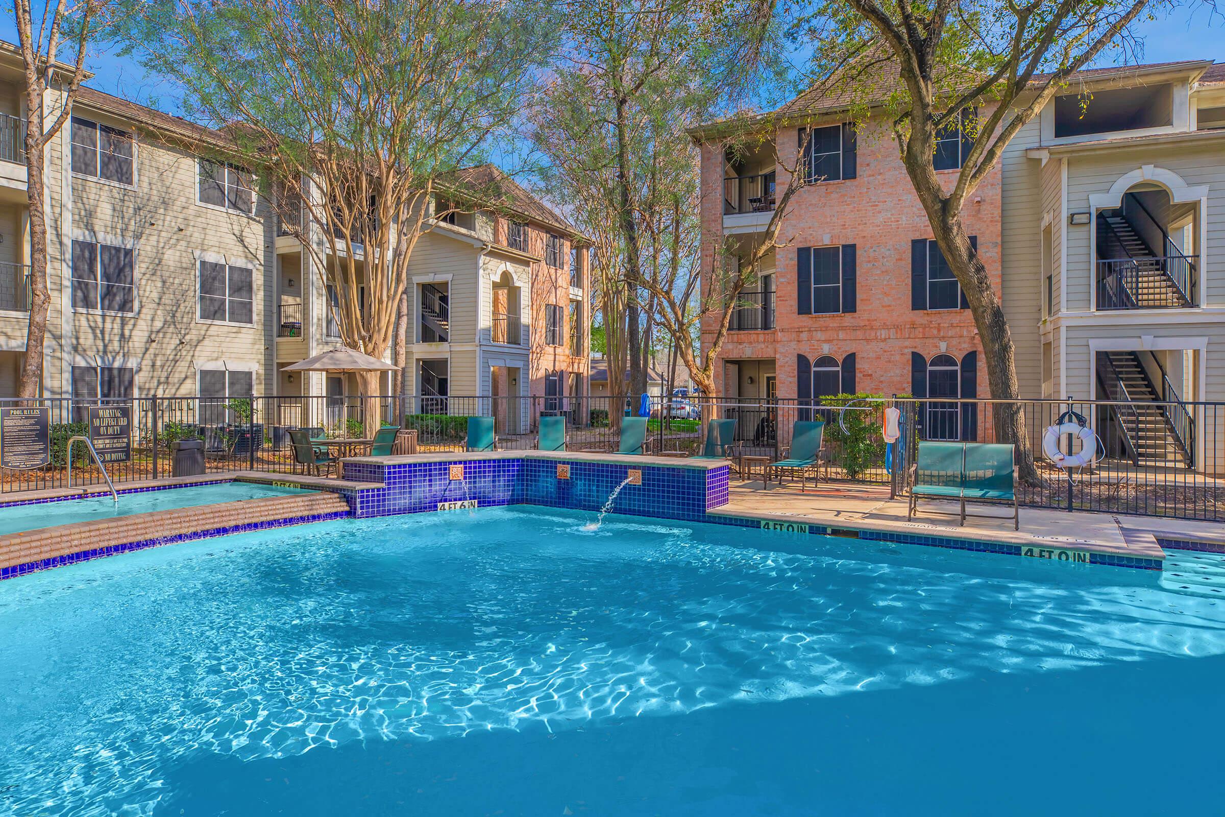 A clear blue swimming pool surrounded by lounge chairs, with several apartment buildings in the background. The area is shaded by trees, suggesting a relaxing outdoor space. The architecture features a mix of brick and siding, typical of modern apartment complexes.