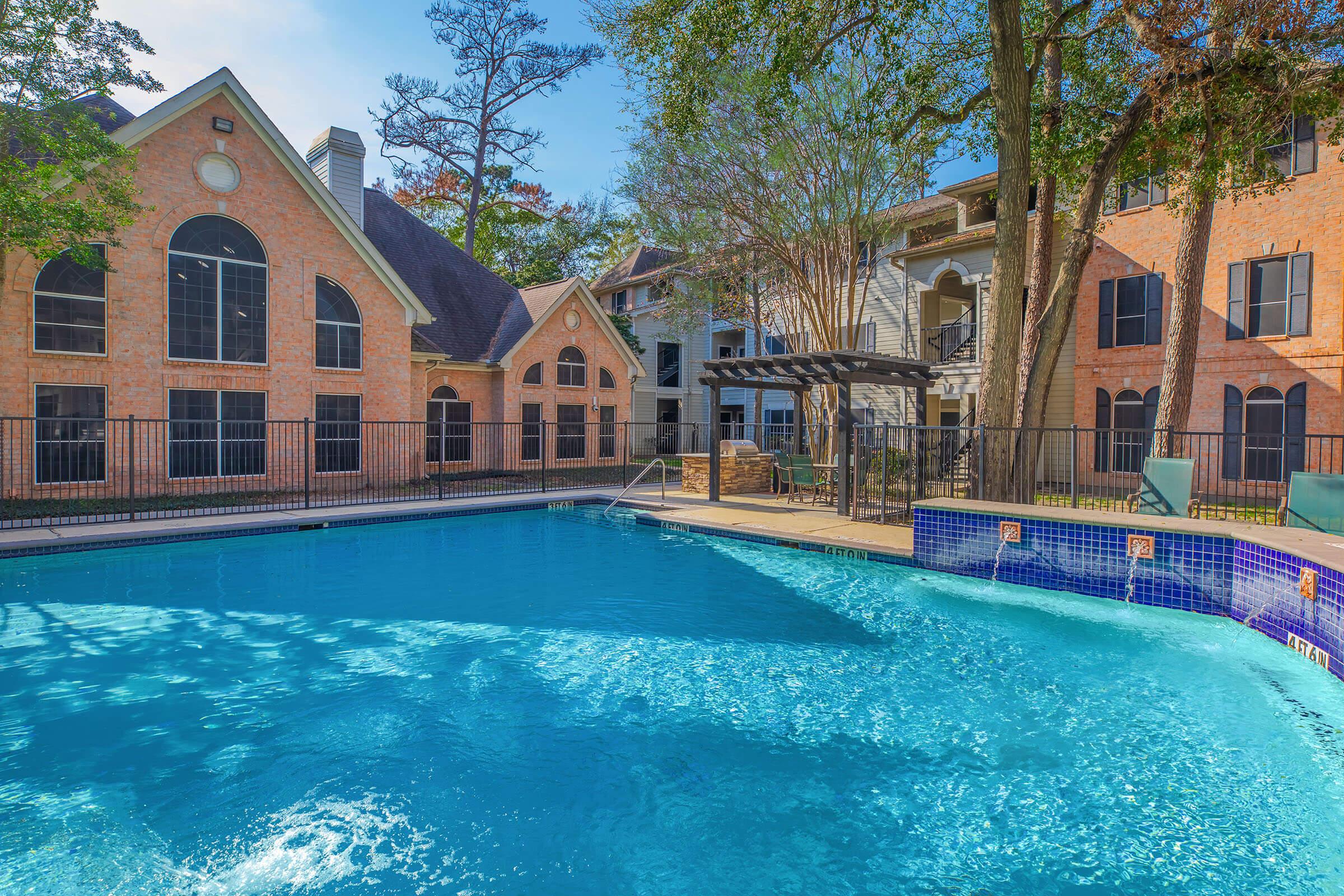 A sparkling blue swimming pool surrounded by brick buildings with large windows and a pergola. Lush greenery and trees are visible in the background, creating a relaxing atmosphere. The pool area features lounge chairs and is well-maintained, suggesting a residential or leisure property.