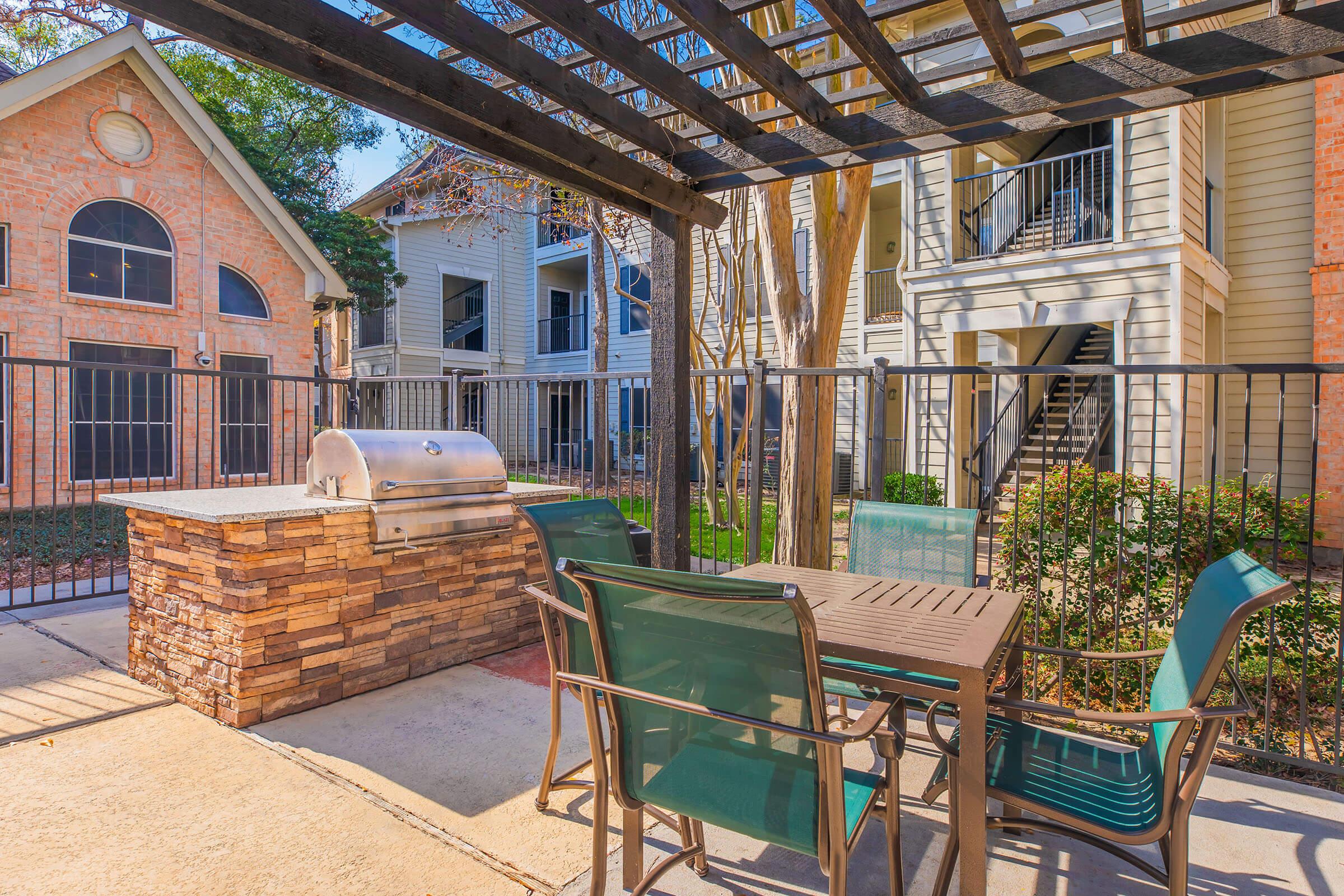 Outdoor grilling area featuring a stainless steel barbecue grill, stone accents, and a table with four green chairs under a wooden pergola. Surrounding the area are landscaped shrubs and nearby apartment buildings, creating a cozy and inviting space for outdoor dining and gatherings.
