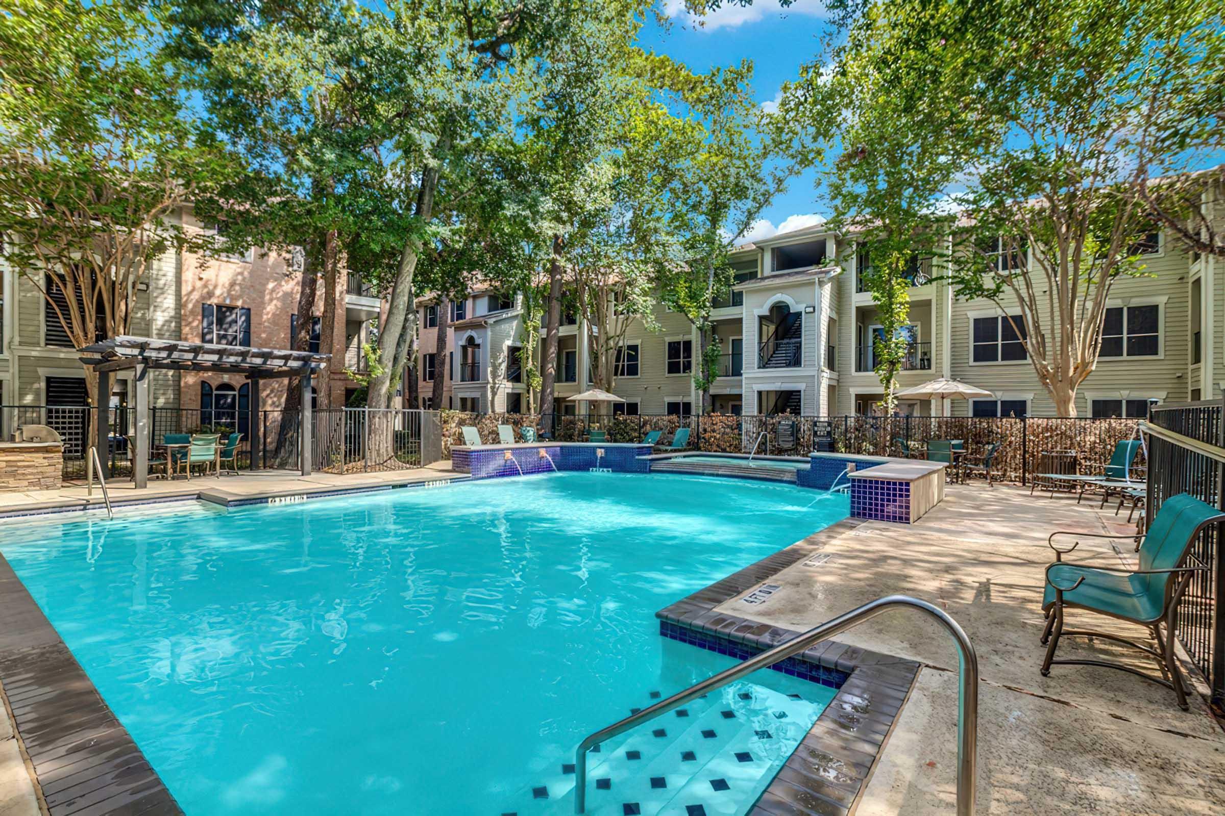 A vibrant outdoor swimming pool surrounded by lush trees and clear blue skies. Lounge chairs and patio furniture are set up around the pool area, with an apartment complex in the background featuring multiple stories and outdoor seating options. A serene, inviting atmosphere perfect for relaxation.