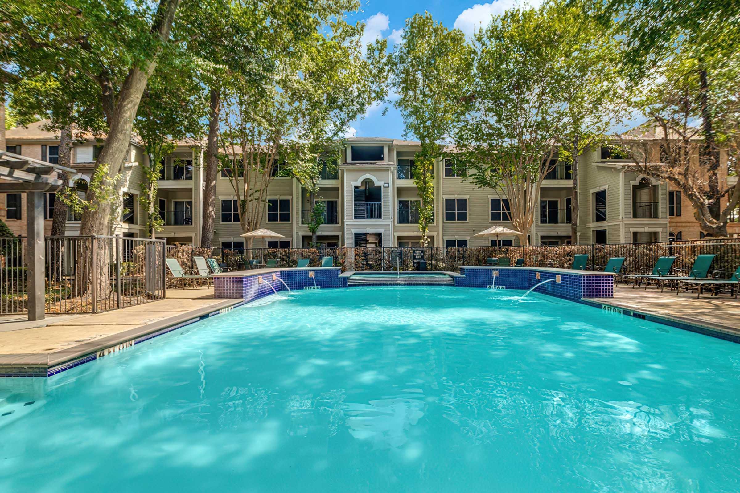 A clear blue swimming pool surrounded by lush greenery and tall trees. In the background, there are several apartment buildings with balconies, and lounge chairs line the poolside. The scene is bright and inviting, suggesting a relaxing outdoor space.
