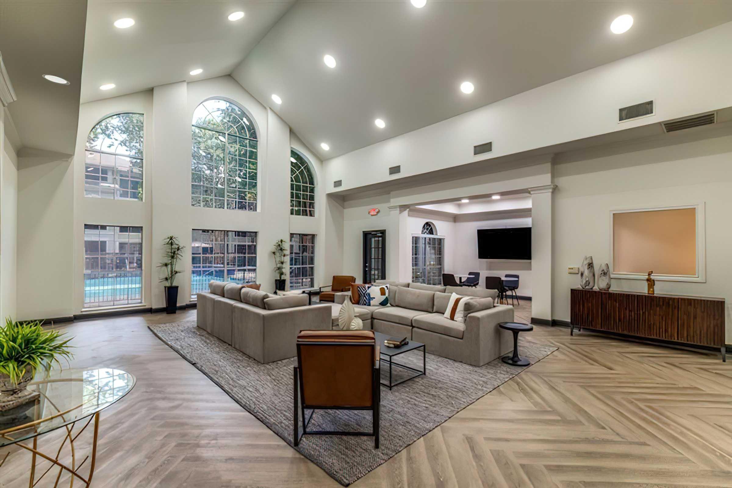 A spacious, modern living area featuring large arched windows with natural light, a comfortable sectional sofa, and stylish furnishings. The room has a warm color palette with wood flooring and a small table. A view of a pool area is visible through the windows, adding to the inviting atmosphere.