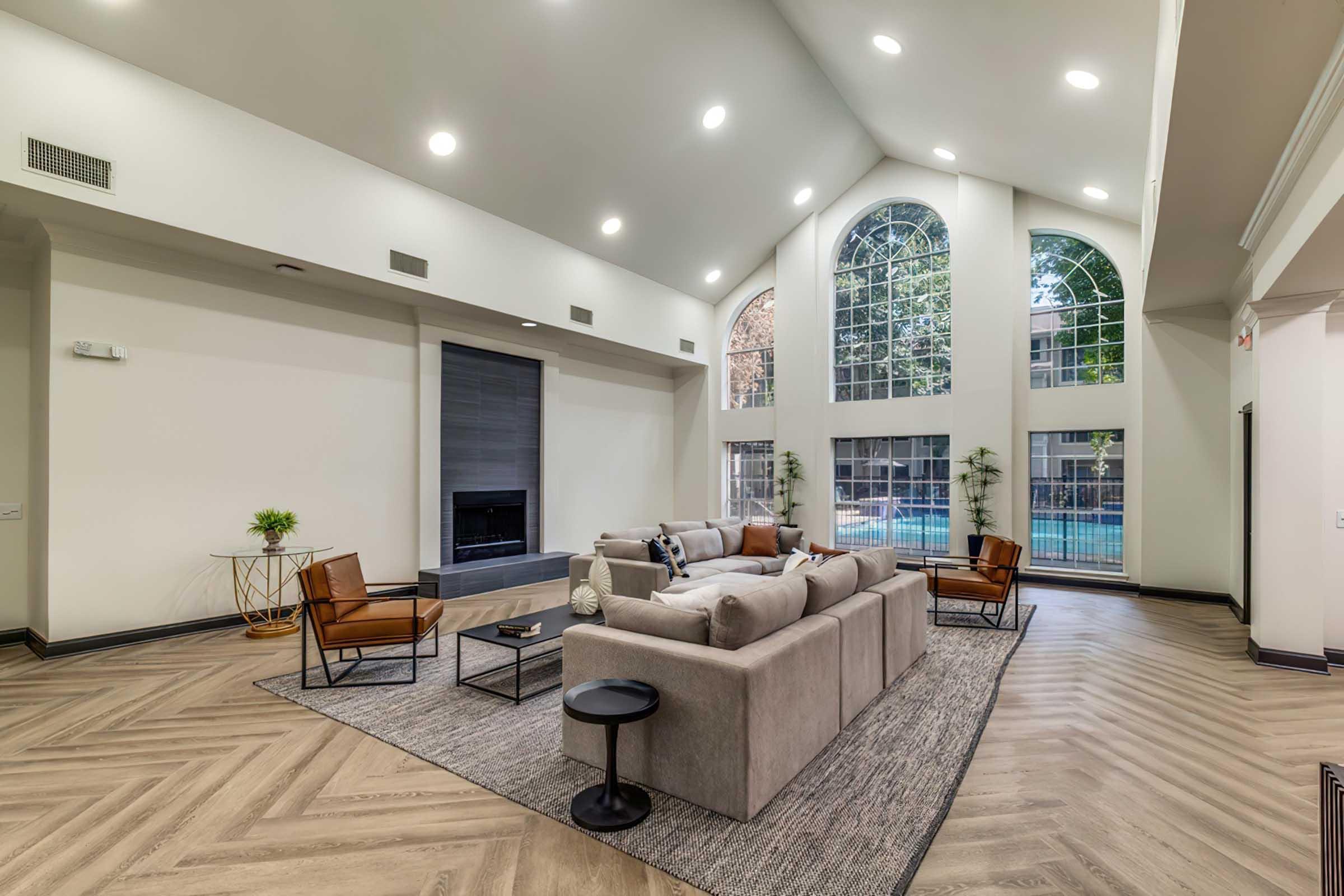 Modern living room with a high ceiling, large windows allowing natural light, and a view of an outdoor pool. The space features a gray sectional sofa, two brown leather chairs, a black side table, and a rug. A minimalist fireplace is on one wall, enhancing the contemporary aesthetic.