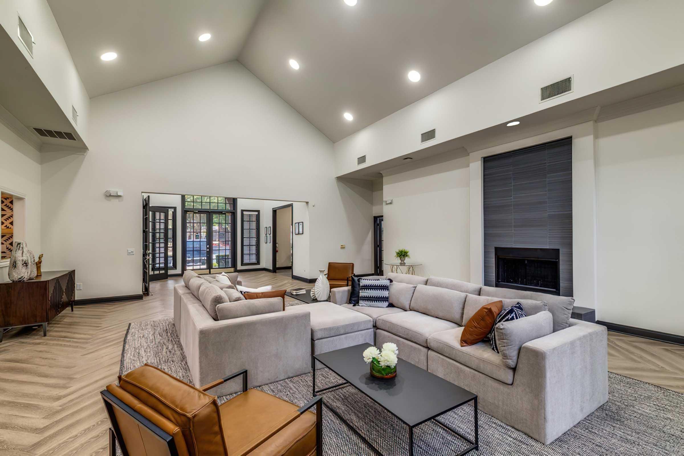 A spacious and modern living room featuring a large gray sectional sofa, a sleek black coffee table, and a decorative plant. The room has high ceilings with recessed lighting, large windows allowing natural light, and a fireplace. There is also an entrance to another room visible in the background.