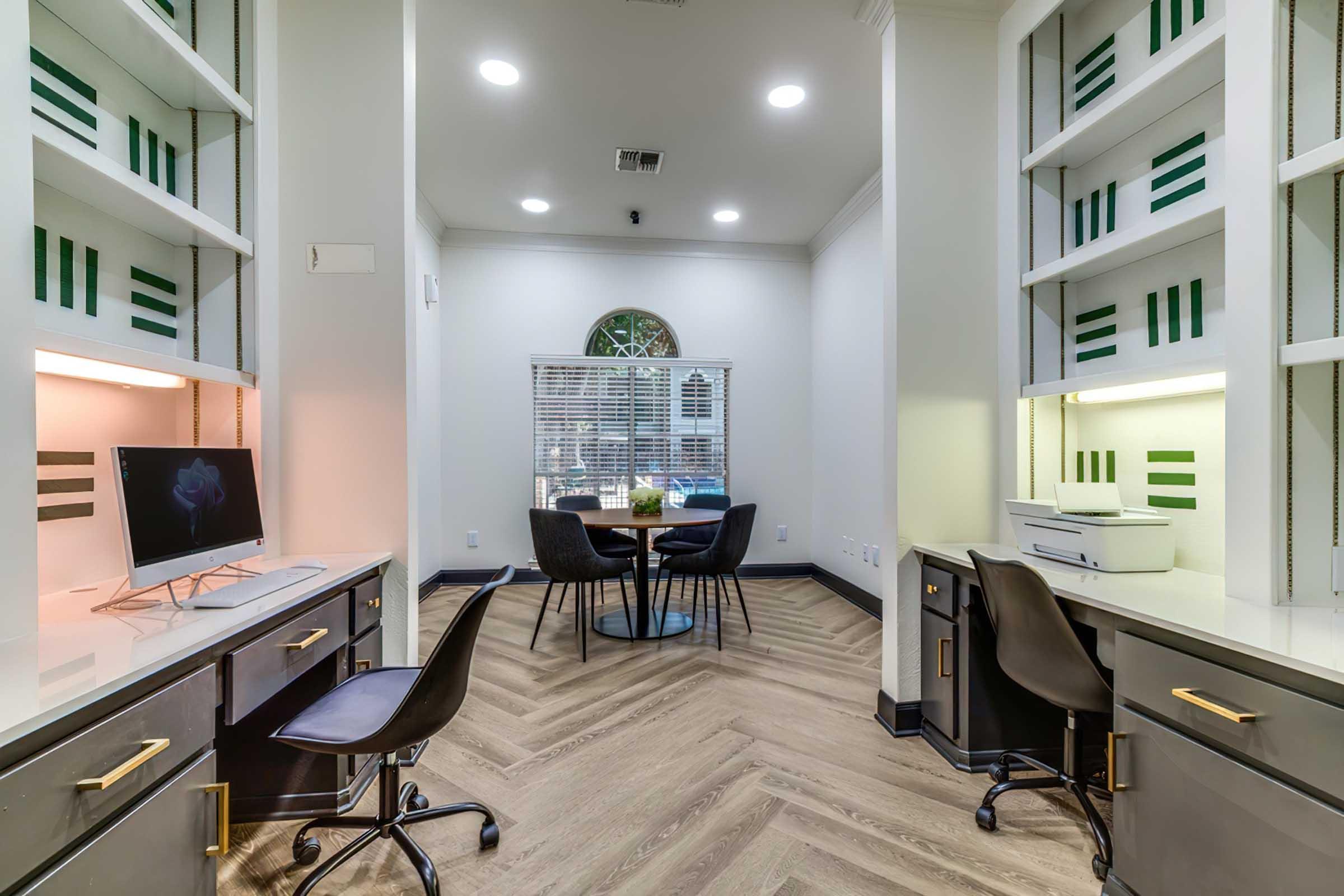 A modern office space featuring two workstations with black chairs, a computer, and a printer. The walls have built-in shelving with green accents. A circular window allows natural light, while a small round table with chairs is visible in the background, enhancing the workspace's functionality and aesthetic.